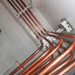 KLIMA DMITRY PETELCZYC - Miedziane rury klimatyzacji, przymocowane do szarej ściany i sufitu za pomocą białych uchwytów. Widok z dołu, instalacja w trakcie montażu.
