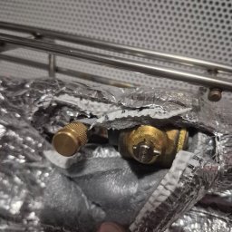 KLIMA DMITRY PETELCZYC - Izolowany zawór klimatyzacji z widocznym elementem regulacyjnym, owinięty folią aluminiową, na tle kratki wentylacyjnej.