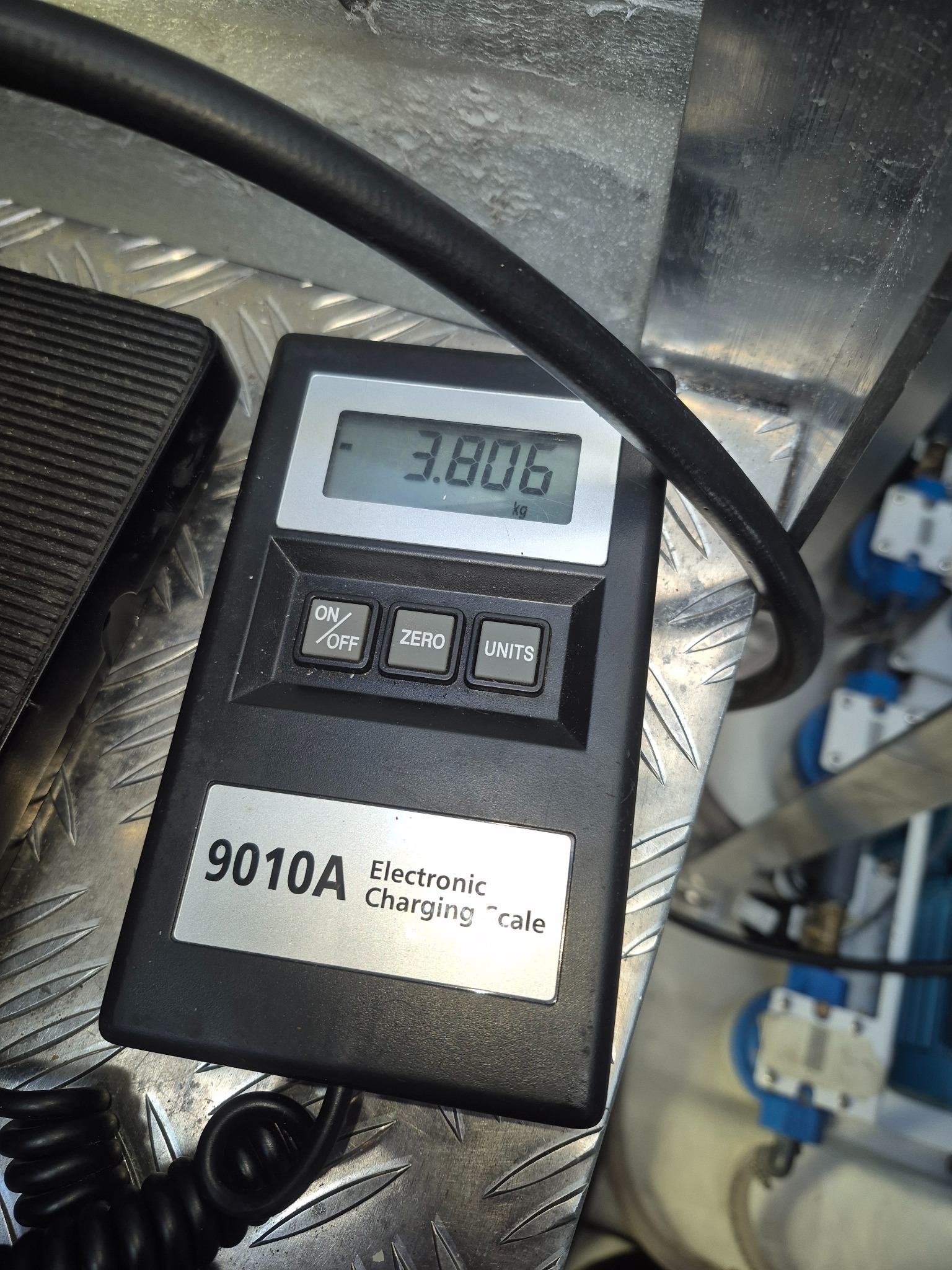 Elektroniczna waga do pomiaru czynnika chłodniczego klimatyzacji, wskazująca 3.806 kg, leżąca na metalowej powierzchni roboczej z widocznymi elementami instalacji.