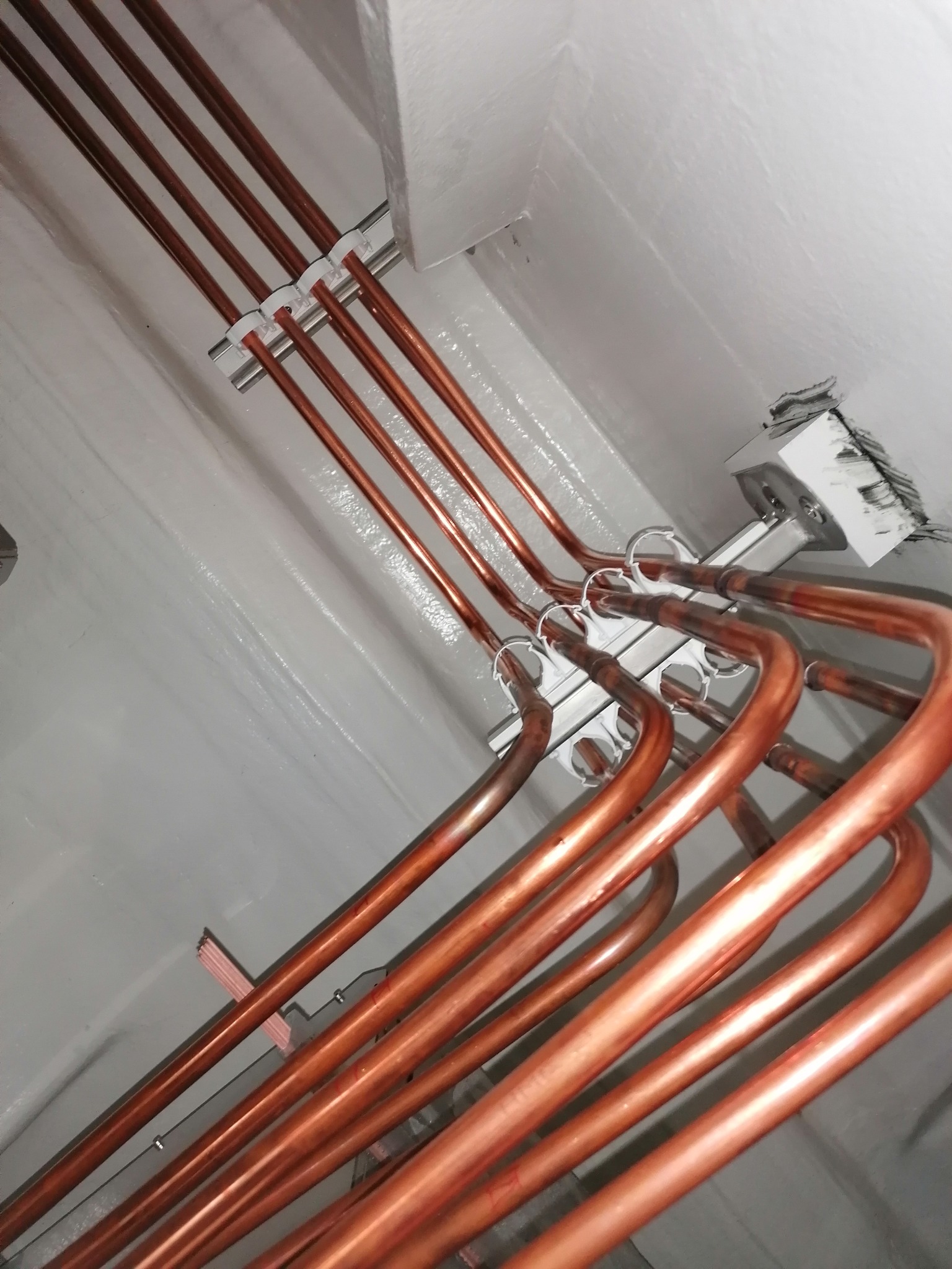 Miedziane rury klimatyzacji, przymocowane do szarej ściany i sufitu za pomocą białych uchwytów. Widok z dołu, instalacja w trakcie montażu.