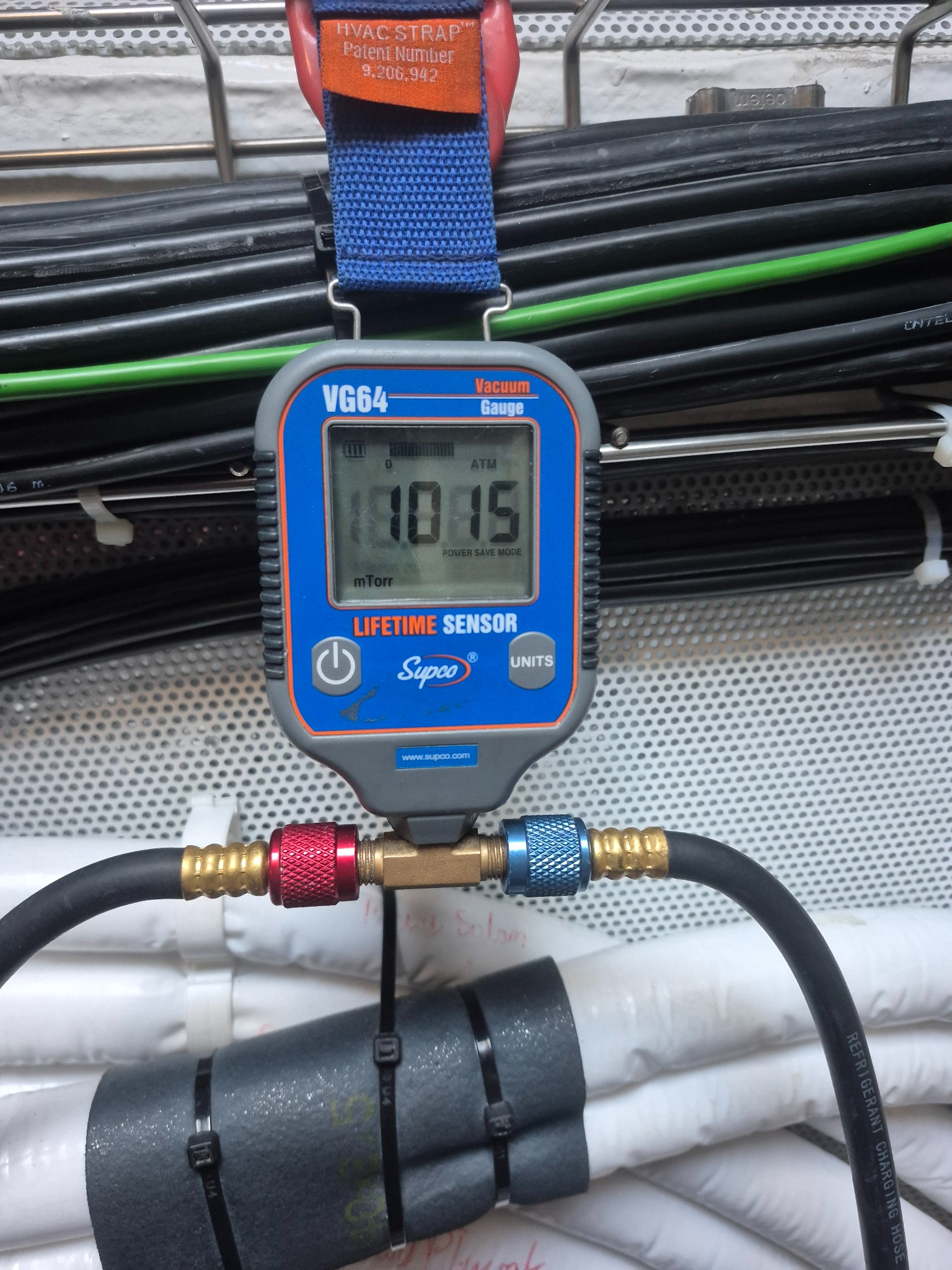 Miernik podciśnienia VG64 Supco, podłączony do przewodów chłodniczych, wskazuje wartość 1015 mTorr. Widoczne przewody elektryczne i izolacja termiczna.