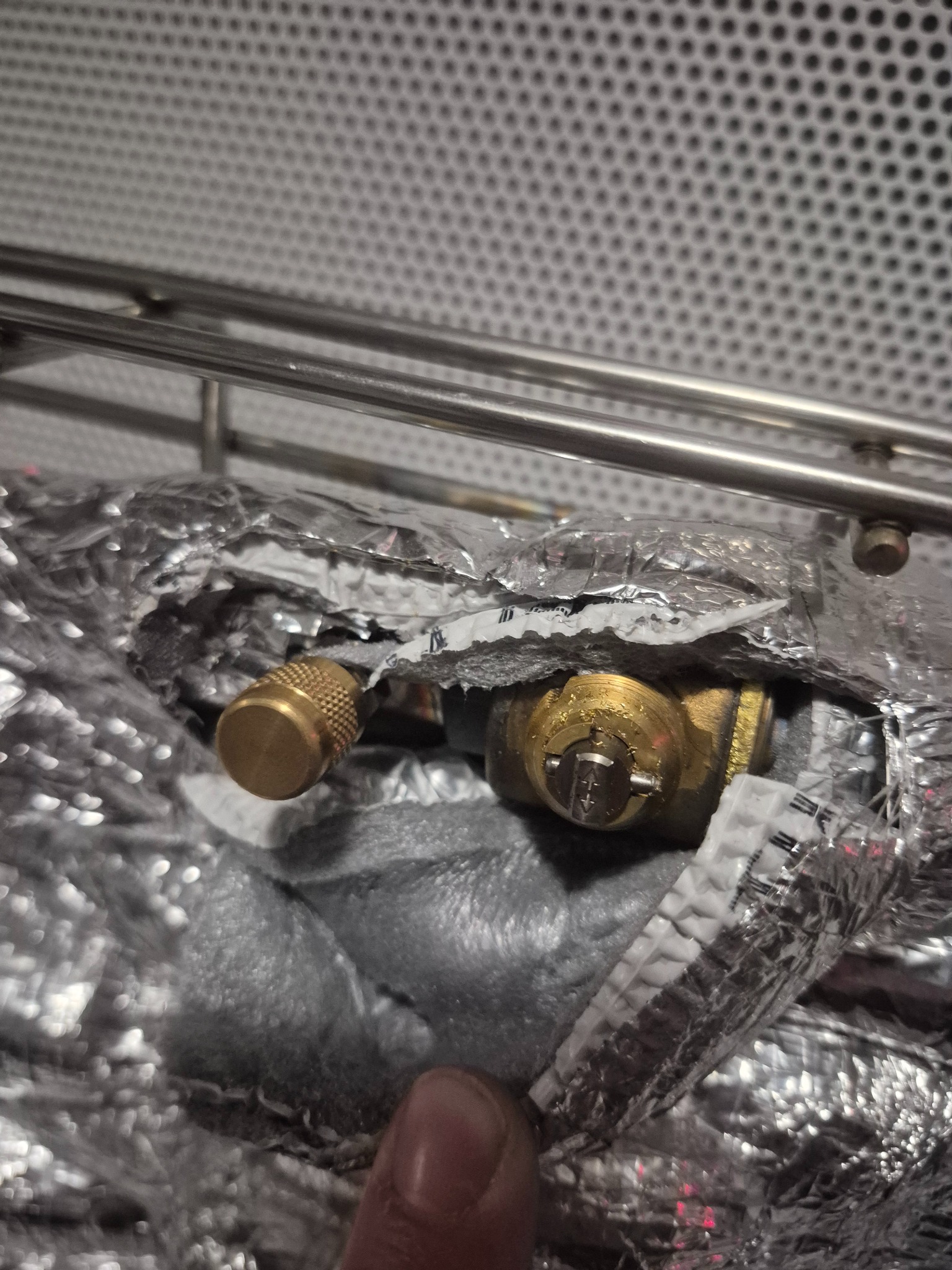 Izolowany zawór klimatyzacji z widocznym elementem regulacyjnym, owinięty folią aluminiową, na tle kratki wentylacyjnej.