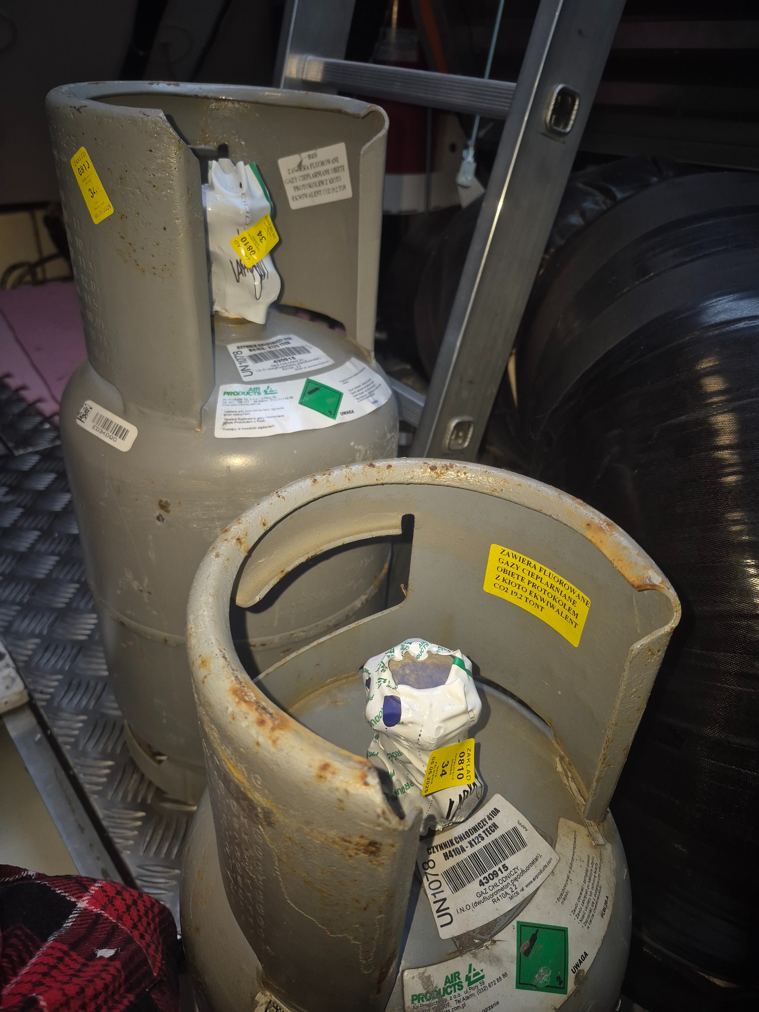 Dwie szare butle z gazem chłodniczym R410A z żółtymi etykietami i naklejkami informacyjnymi, ustawione obok siebie w nieokreślonym miejscu pracy.