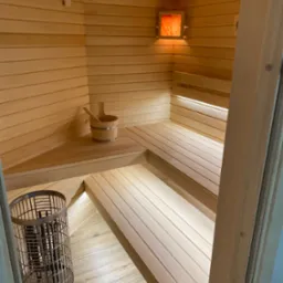 Jasna, drewniana sauna z piecem, ławkami i oświetleniem. Drewniany kubełek z chochlą na półce. Widok z zewnątrz, przez otwarte drzwi.