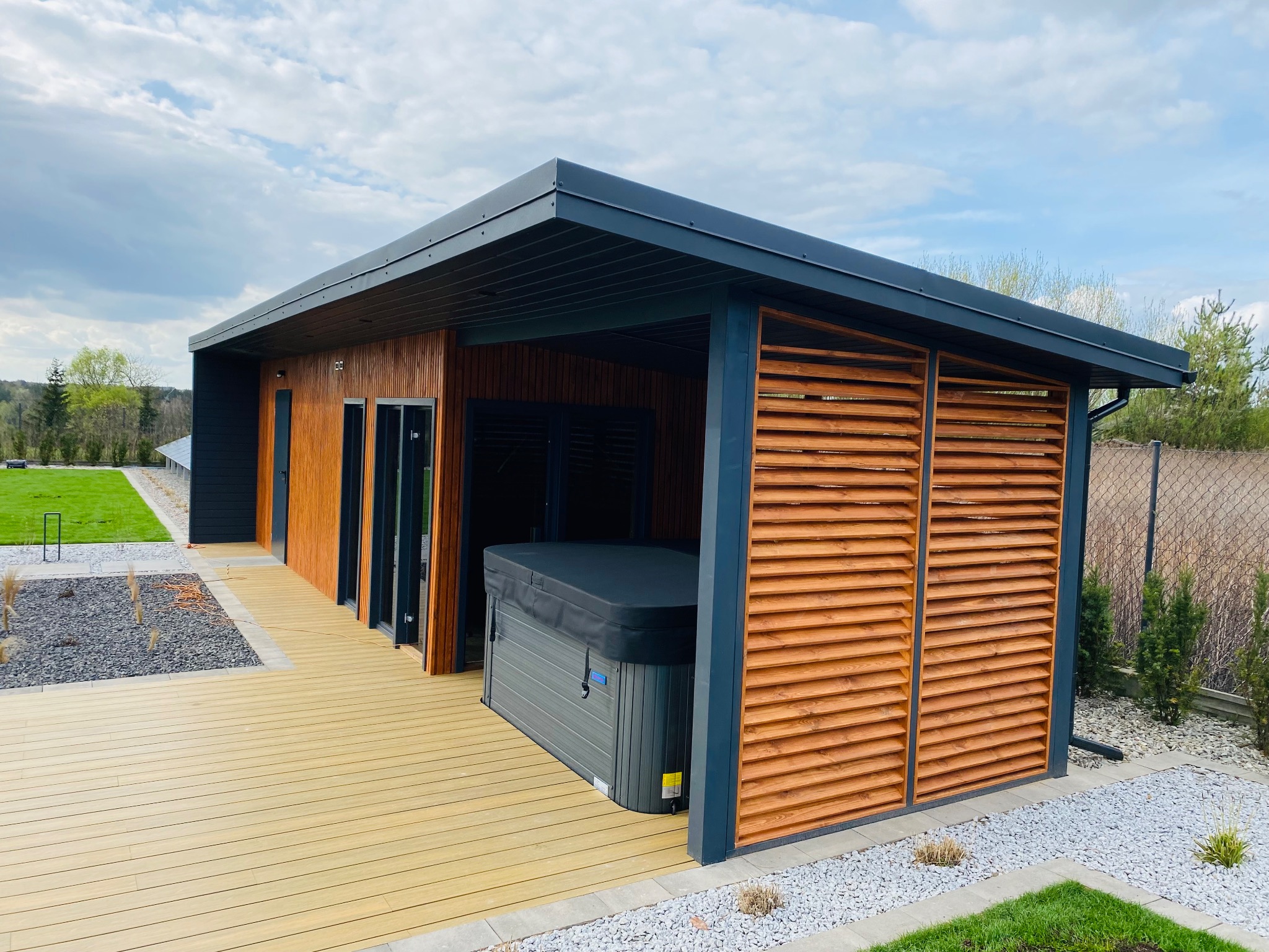 Nowoczesna konstrukcja z drewna i kompozytu z zadaszeniem, osłaniająca jacuzzi na tarasie, z widokiem na zadbany ogród i błękitne niebo.