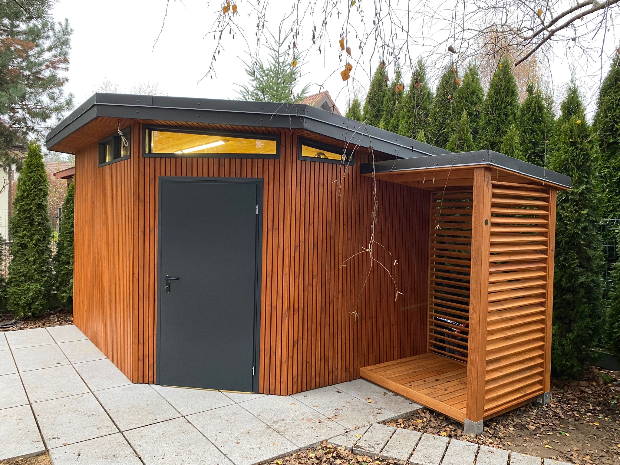 Nowoczesna, drewniana altana ogrodowa z ciemnoszarymi drzwiami i zadaszeniem, połączona z ażurową konstrukcją na drewno, na tle zielonych tuj i szarej kostki brukowej.