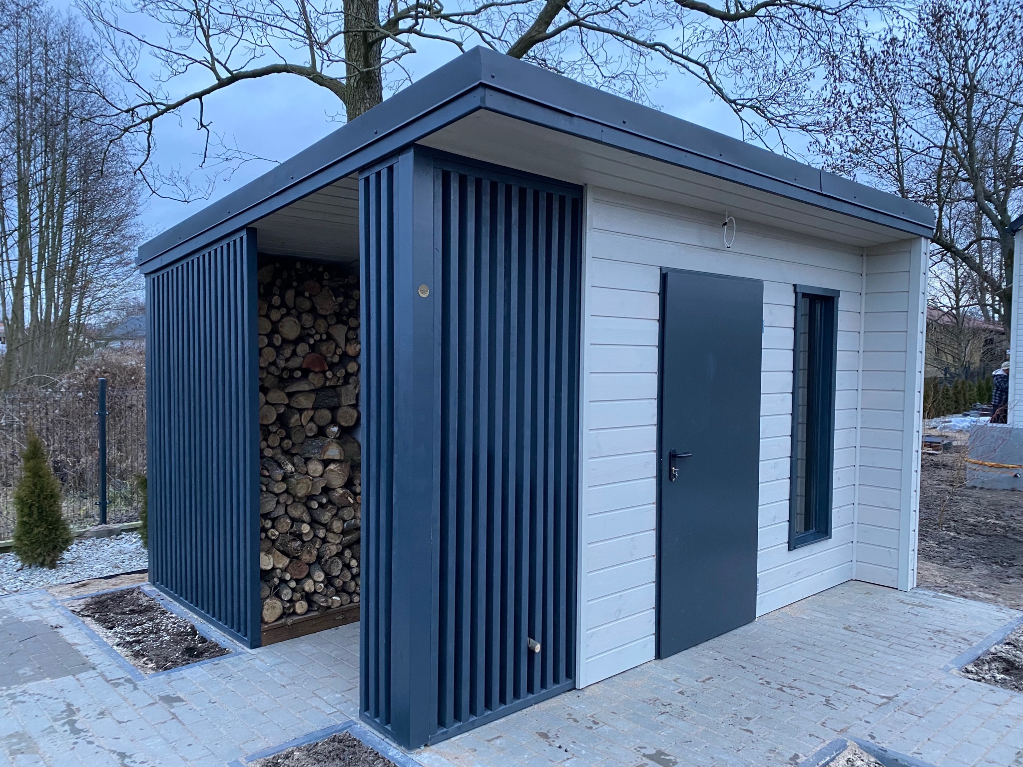 Nowoczesna konstrukcja z drewna i metalu w kolorze szaro-białym, z otwartą przestrzenią na drewno opałowe i zamkniętym pomieszczeniem z drzwiami i oknem, na tle drzew i kostki brukowej.