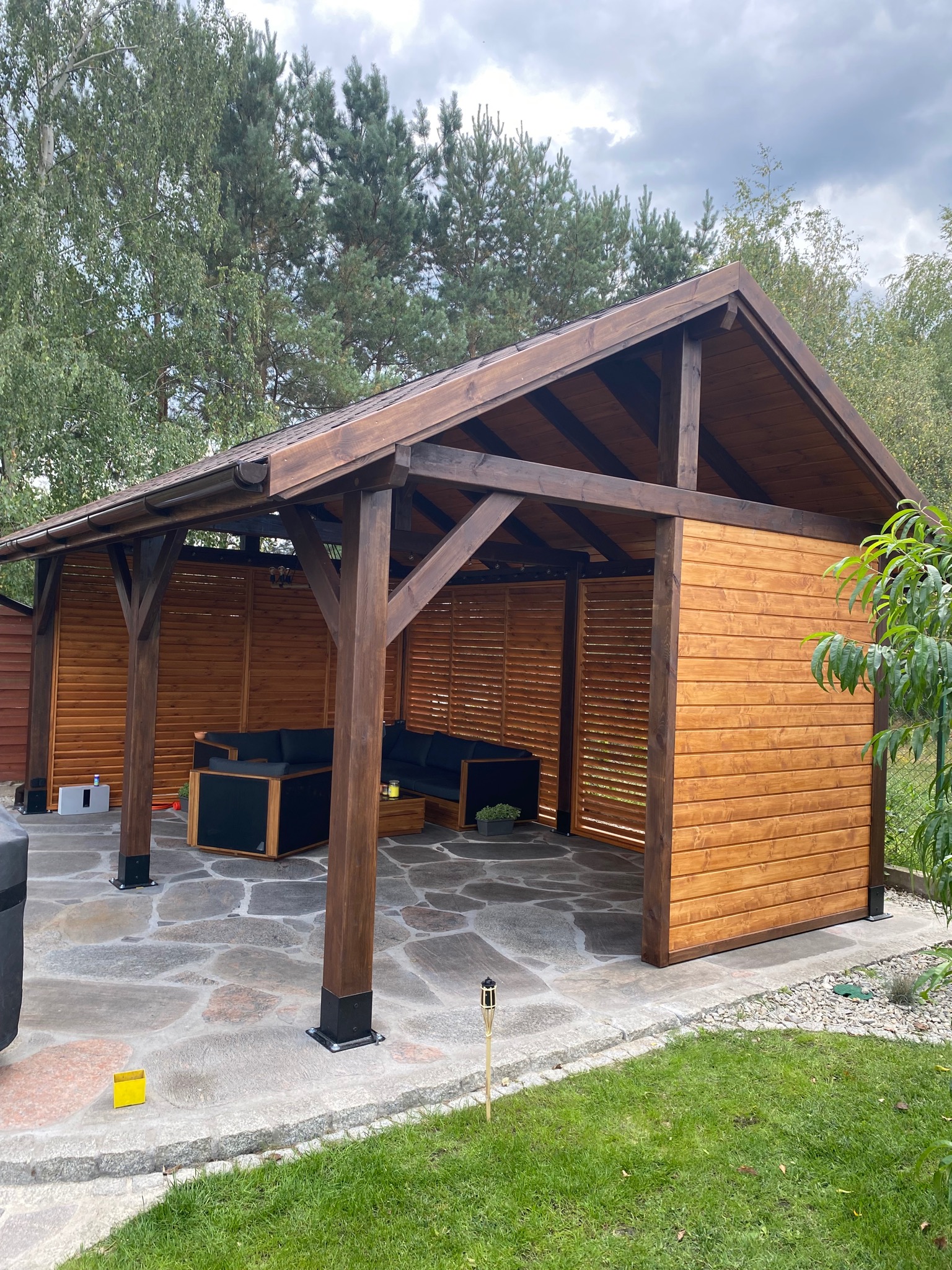 Zadaszona konstrukcja z drewna z ażurowymi ścianami, wyposażona w ciemne meble ogrodowe, umieszczona na kamiennym tarasie z otaczającą zielenią.