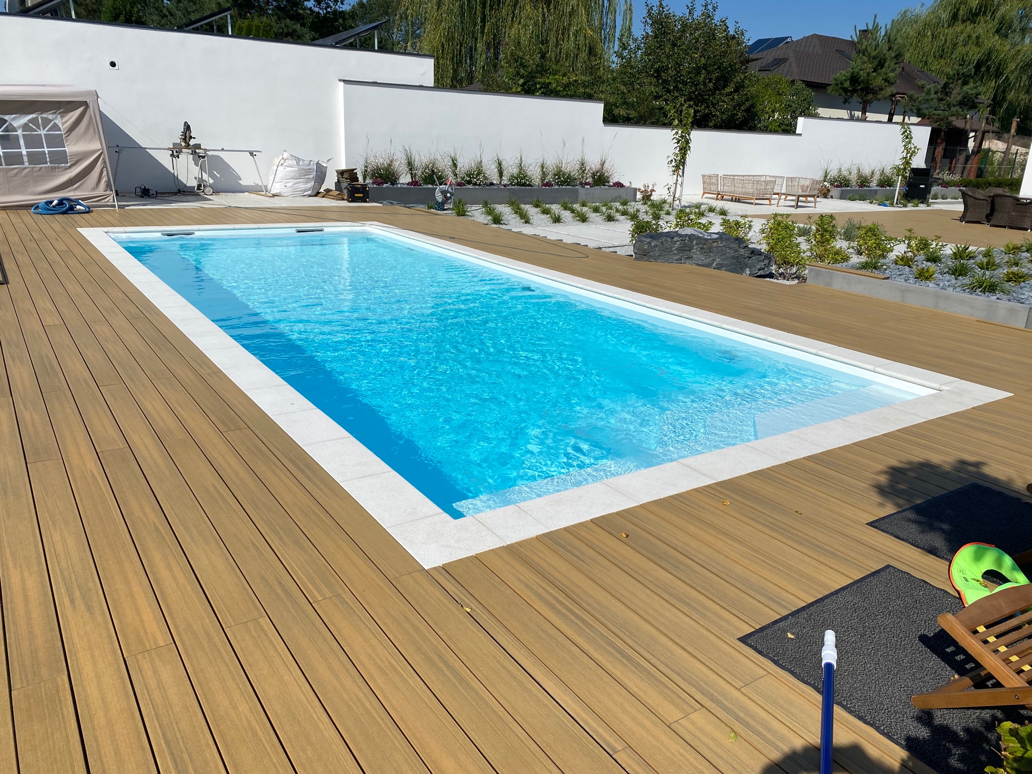 Nowoczesny basen z błękitną wodą otoczony tarasem z desek kompozytowych, widok na biały mur z nasadzeniami i elementy wyposażenia ogrodu.