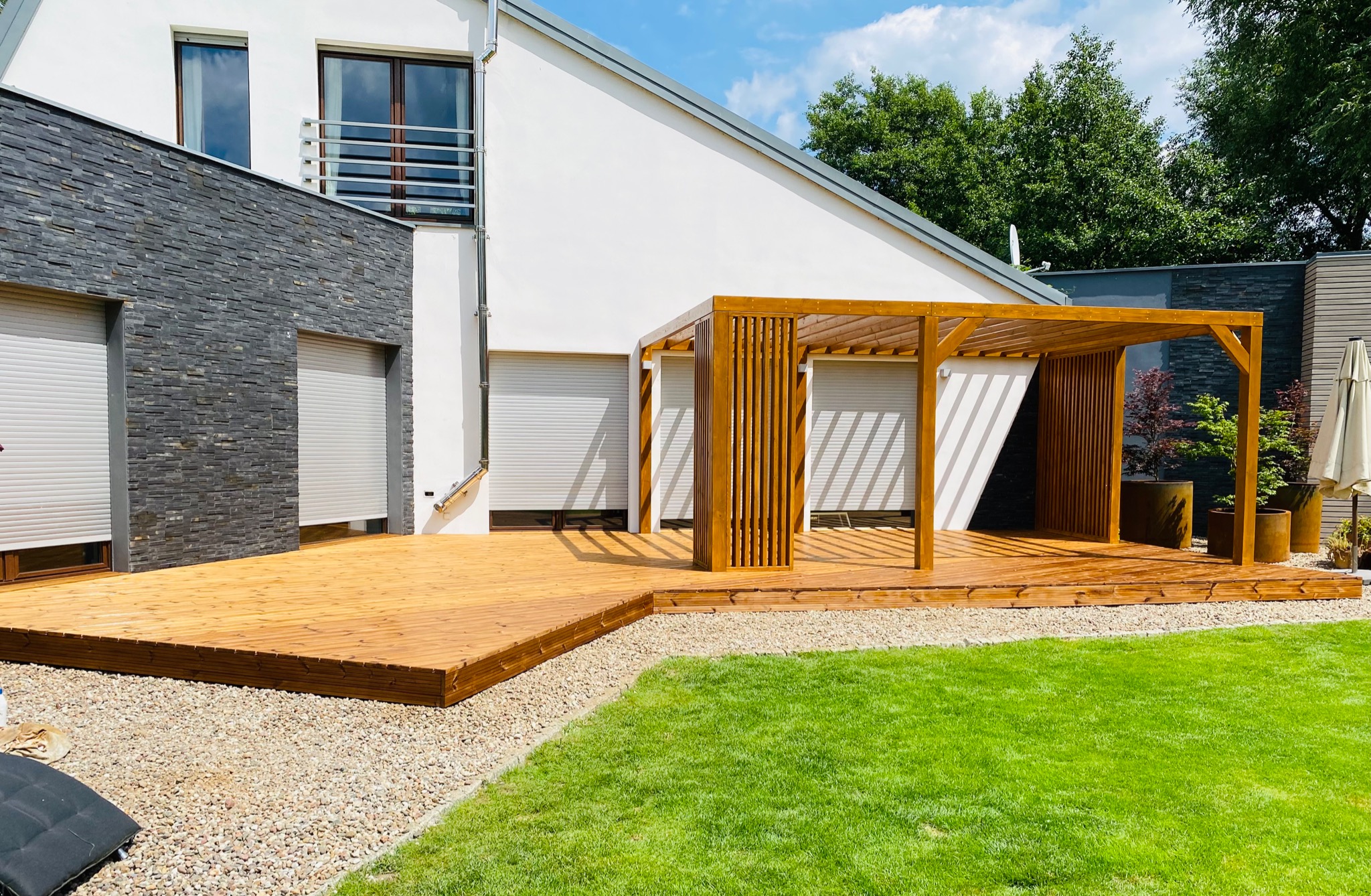 Nowoczesna drewniana konstrukcja zadaszenia tarasu z pionowymi listwami, przylegająca do jasnego domu z elementami kamiennej elewacji i drewnianym tarasem, otoczona zielonym trawnikiem i żwirowym...