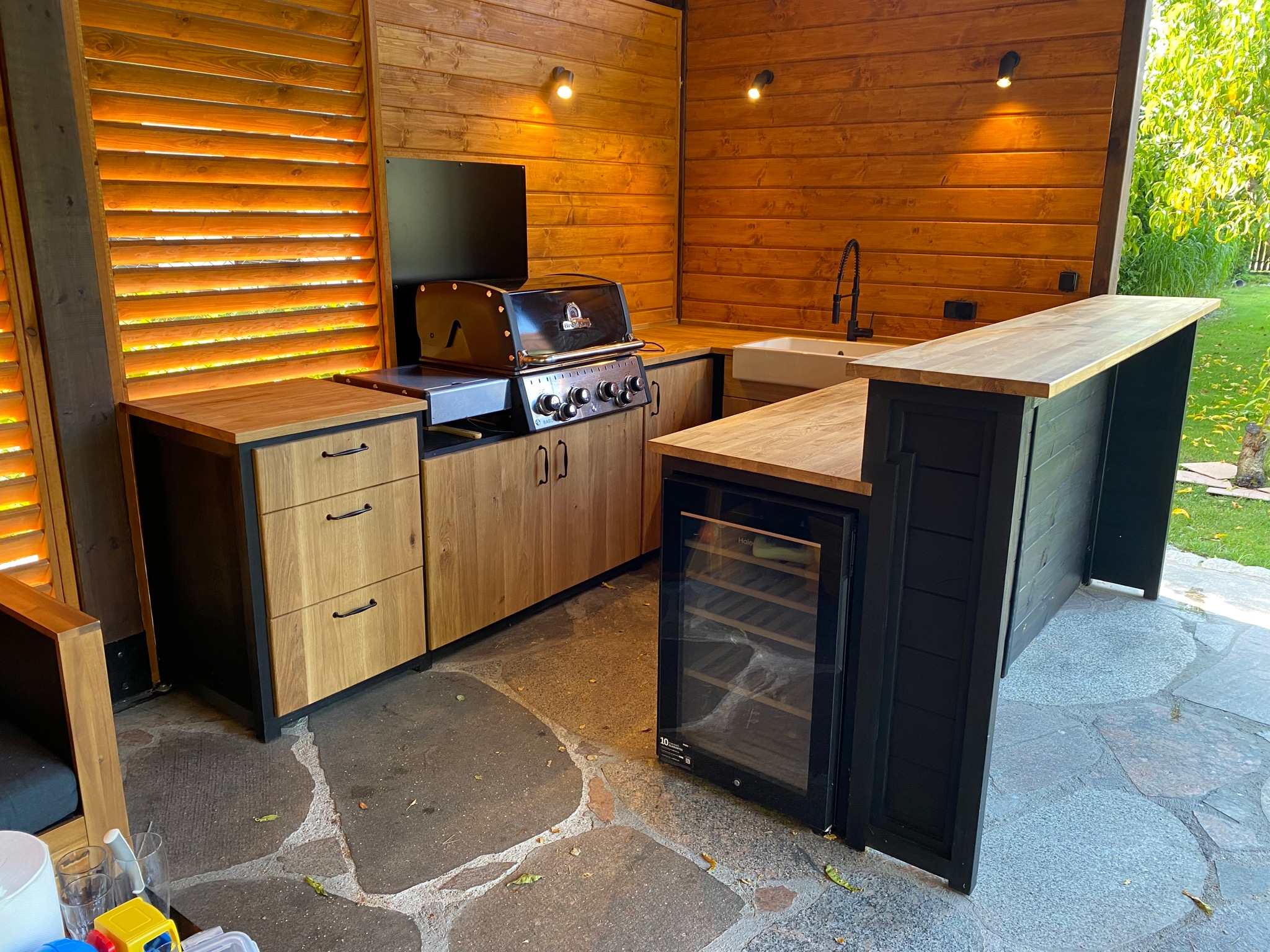 Zadaszona letnia kuchnia z drewnianymi ścianami, blatem, szafkami i grillem, oświetlona punktowymi lampami, z kamienną podłogą i widokiem na zieleń ogrodu.