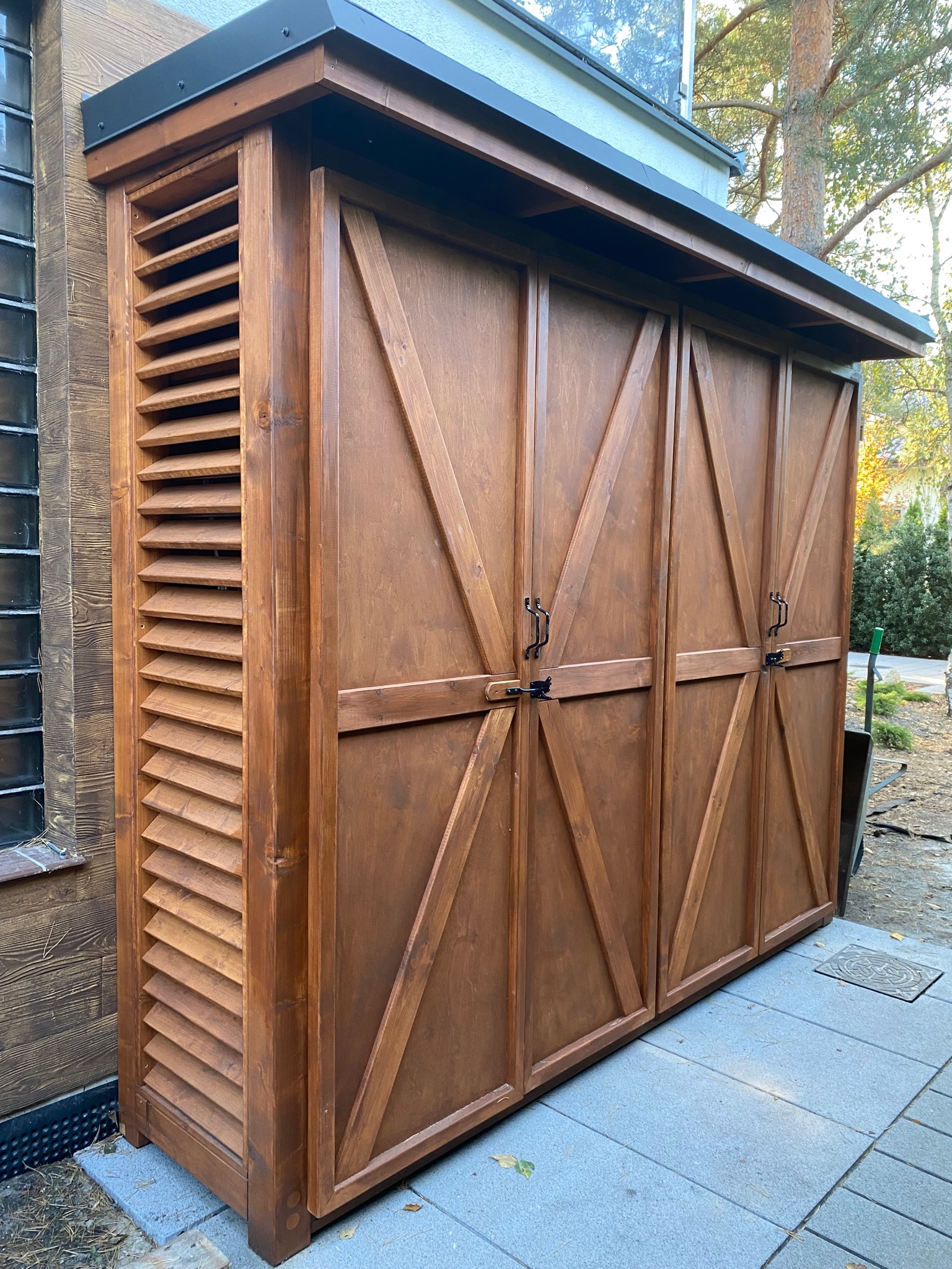 Drewniana konstrukcja z zamykanymi drzwiami i lamelkową ścianą boczną, prawdopodobnie schowek na narzędzia lub akcesoria ogrodowe, umieszczona na zewnątrz budynku.