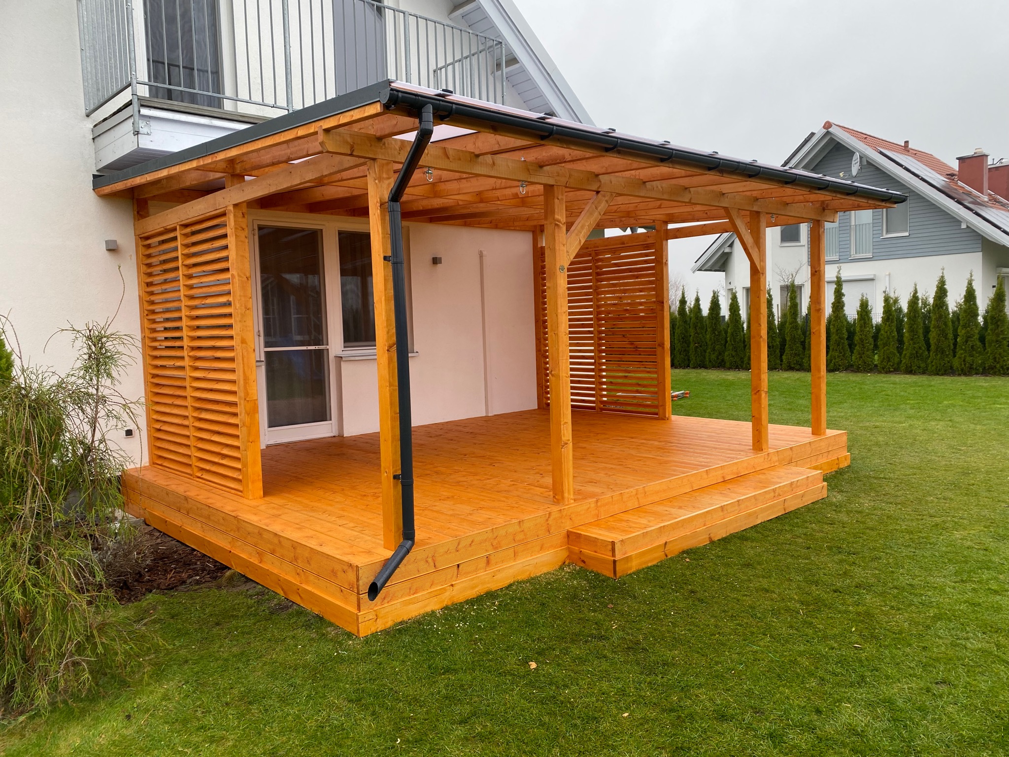 Drewniana pergola z lamelowymi ścianami i tarasem, przylegająca do domu z balkonem, otoczona zielonym trawnikiem i tujami w pochmurny dzień.