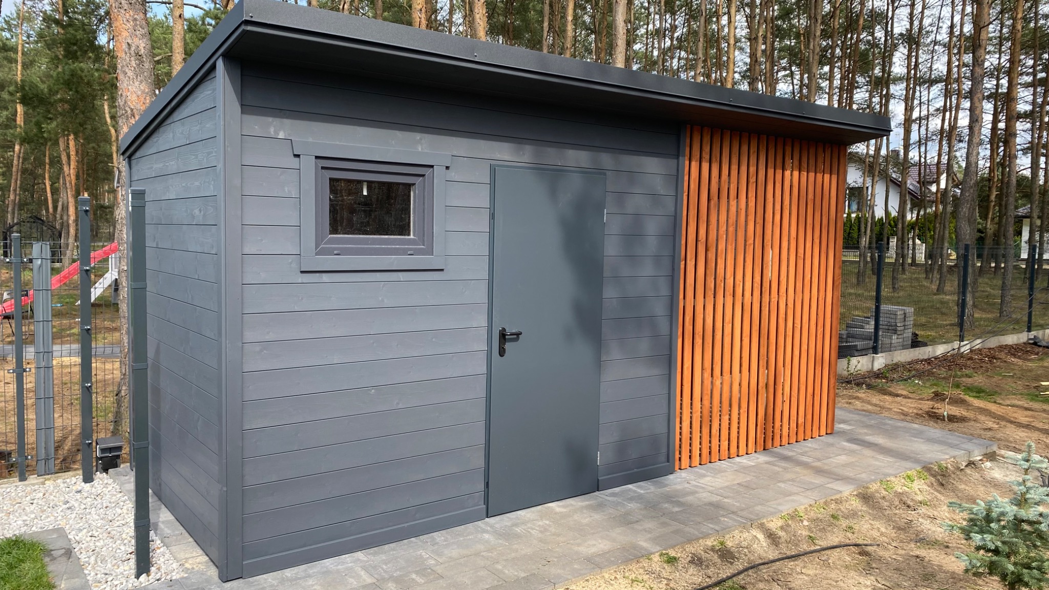 Nowoczesna, szara drewutnia z oknem i pionowymi, drewnianymi panelami na tle leśnego krajobrazu. Widoczne szare drzwi i kostka brukowa.