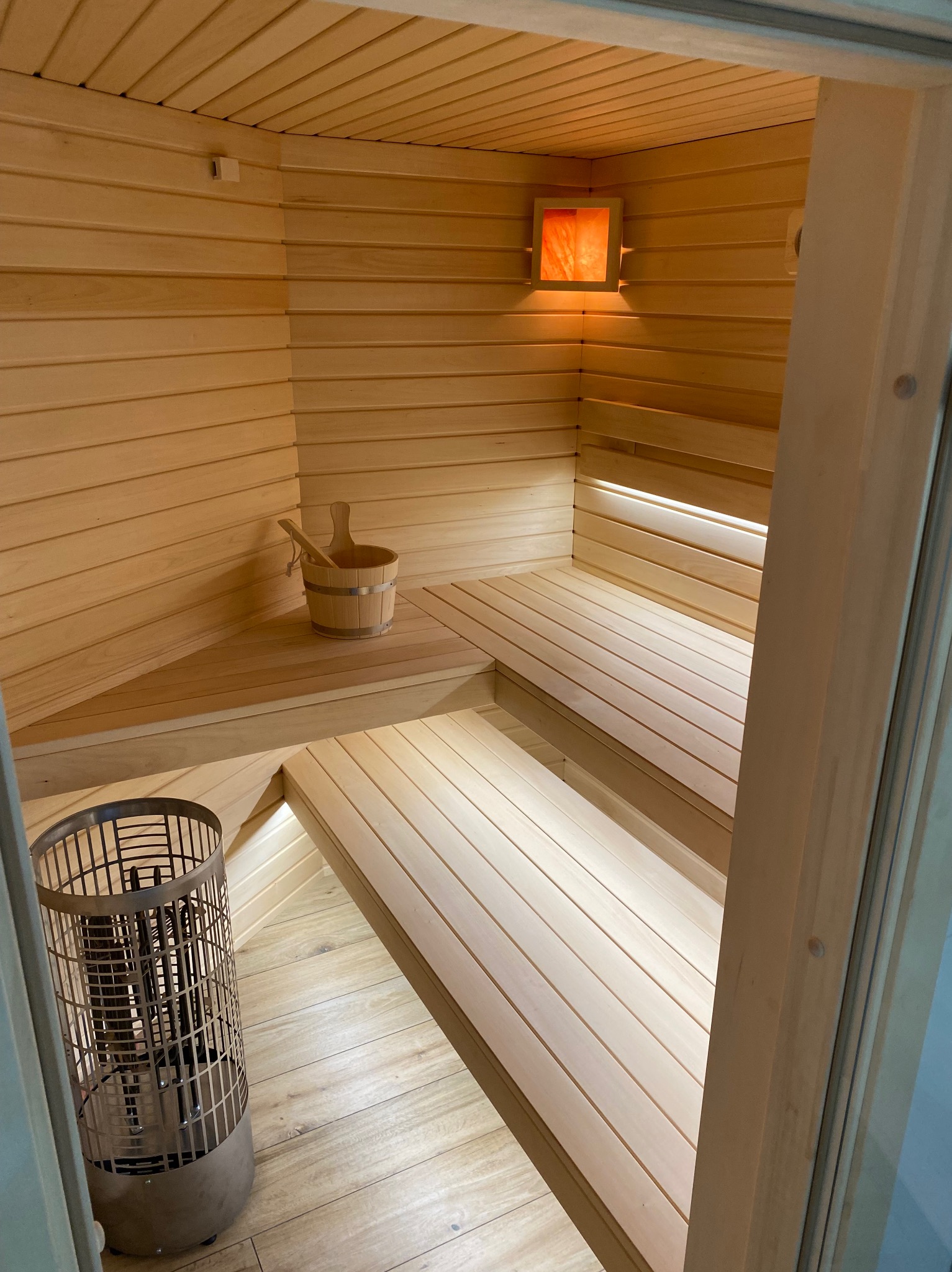 Jasna, drewniana sauna z piecem, ławkami i oświetleniem. Drewniany kubełek z chochlą na półce. Widok z zewnątrz, przez otwarte drzwi.