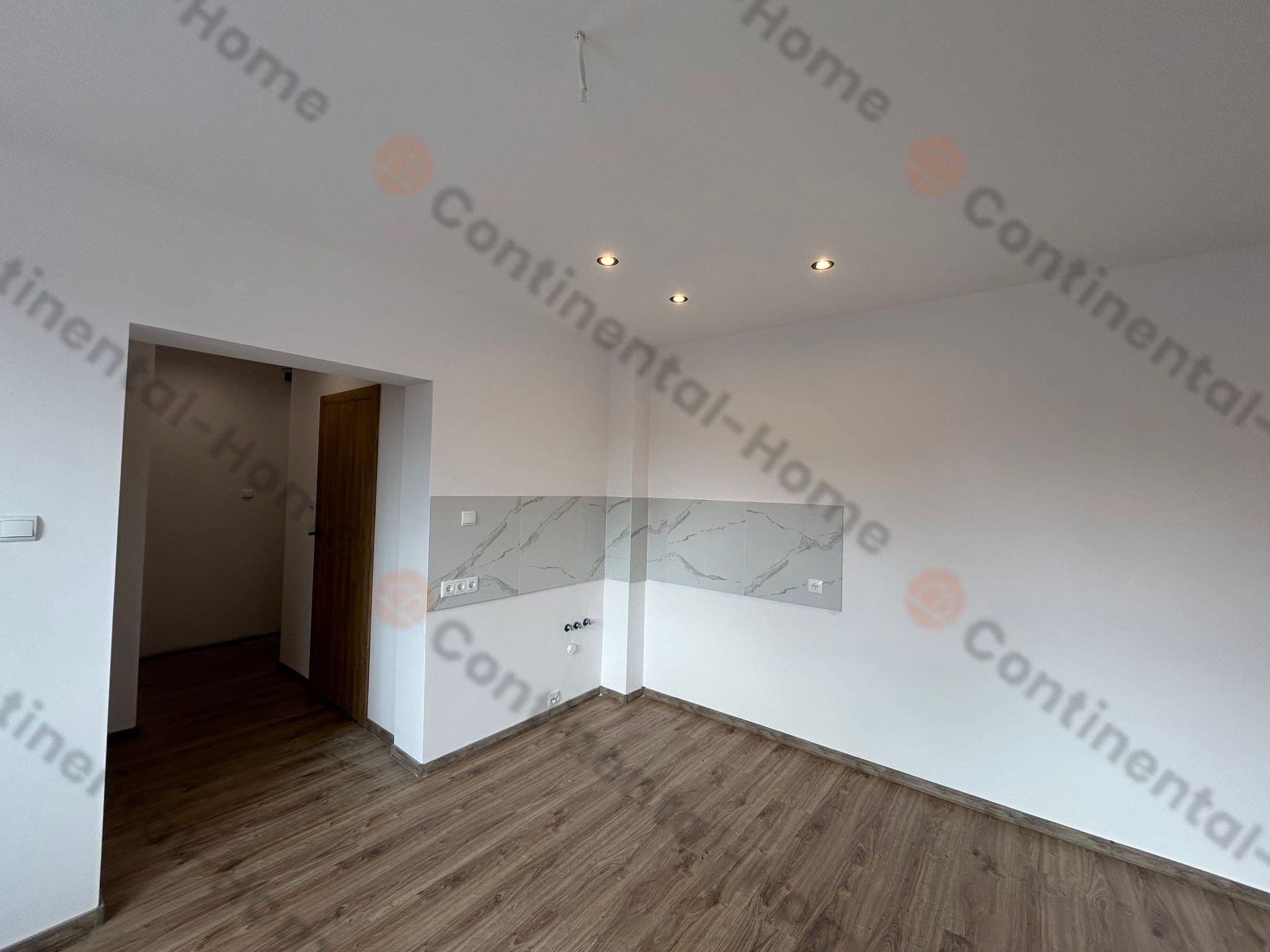 Pusty pokój po remoncie z panelami podłogowymi imitującymi drewno, białymi ścianami, marmurowymi panelami na ścianie, czterema wpuszczonymi punktami świetlnymi w suficie i otwartym przejściem...