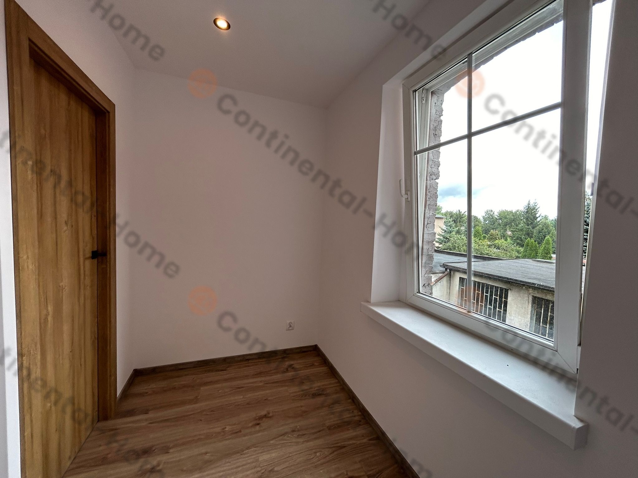 Pusty pokój po odświeżeniu, z drewnianą podłogą, oknem z widokiem na budynek i drzewa oraz drewnianymi drzwiami z czarną klamką.