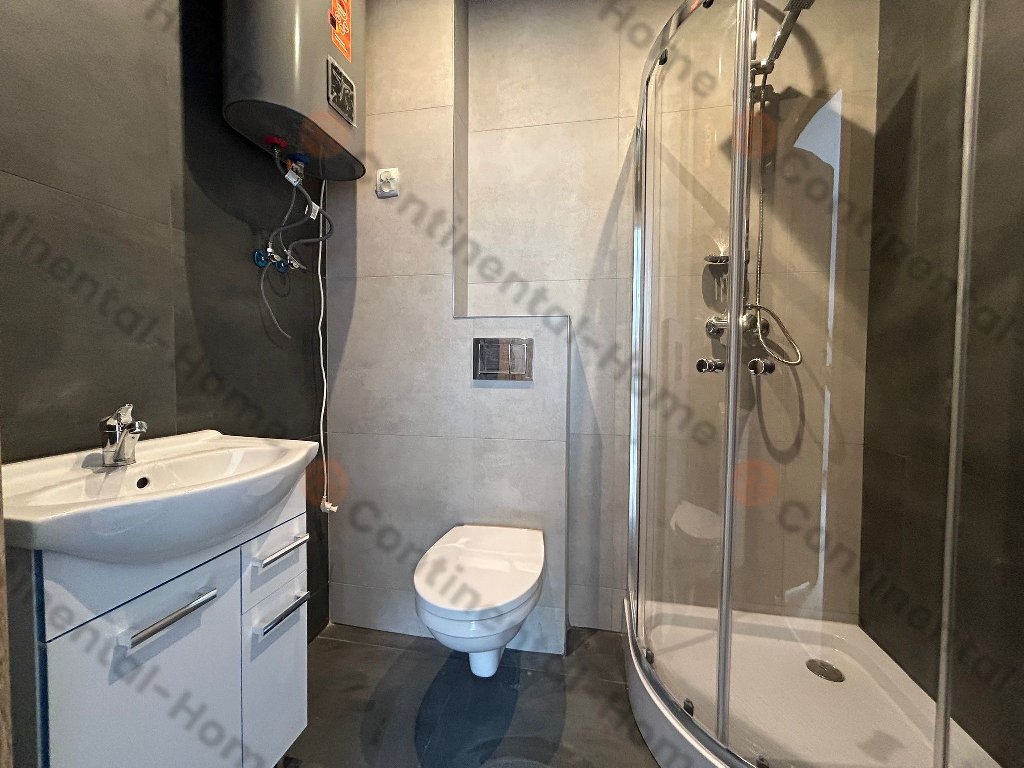 Nowoczesna łazienka z szarymi płytkami, białą umywalką z szafką, toaletą i kabiną prysznicową z hydromasażem oraz bojlerem elektrycznym na ścianie.