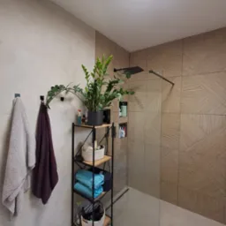 Nowoczesna łazienka z kabiną prysznicową, wykończona płytkami w geometryczny wzór, z rośliną doniczkową na metalowym regale i ręcznikami na wieszakach.