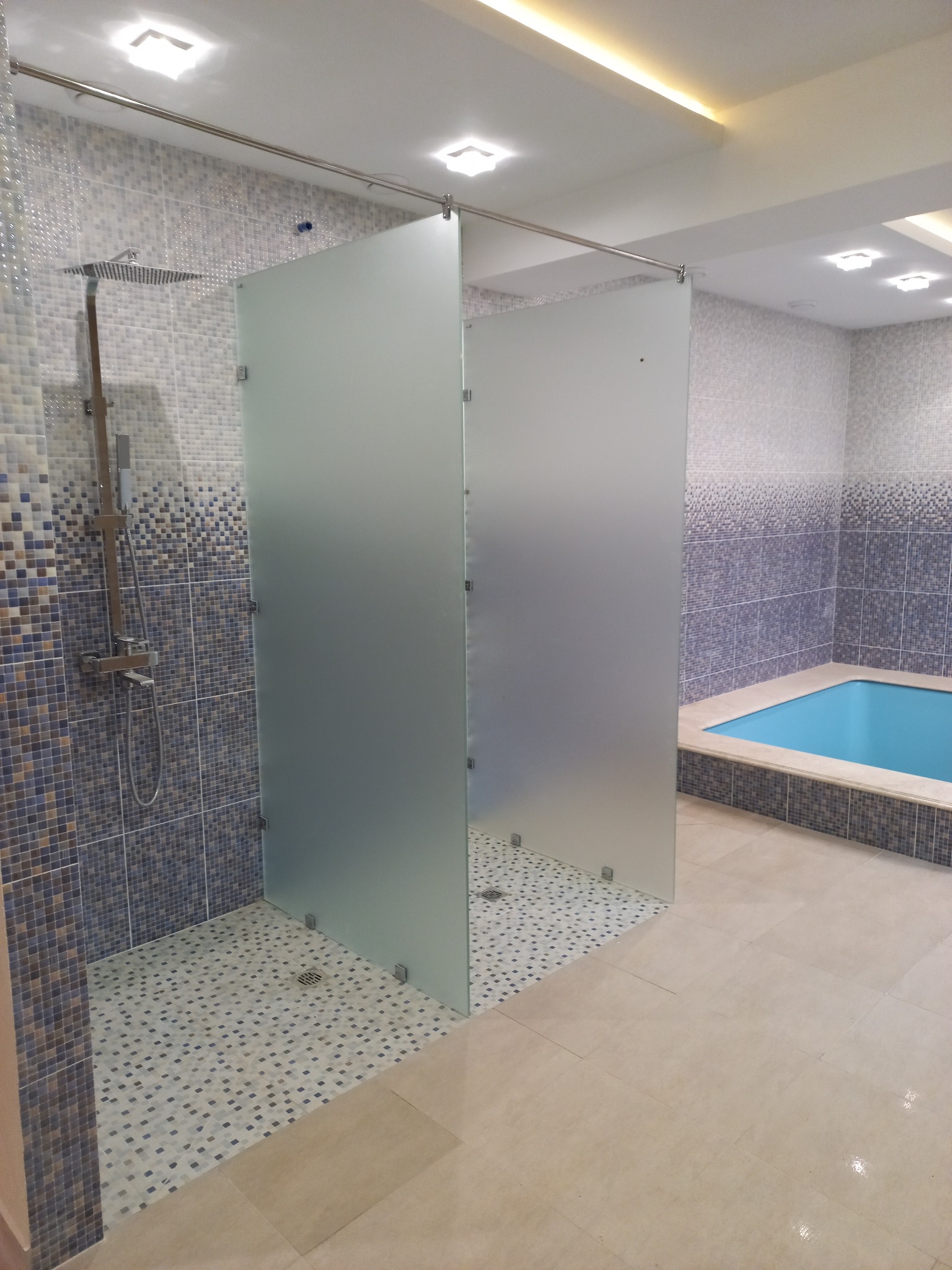 Nowoczesna łazienka z kabiną prysznicową oddzieloną matowymi szklanymi panelami, mozaikowa ściana i podłoga w strefie prysznica, obok mały basen wykończony podobną mozaiką.