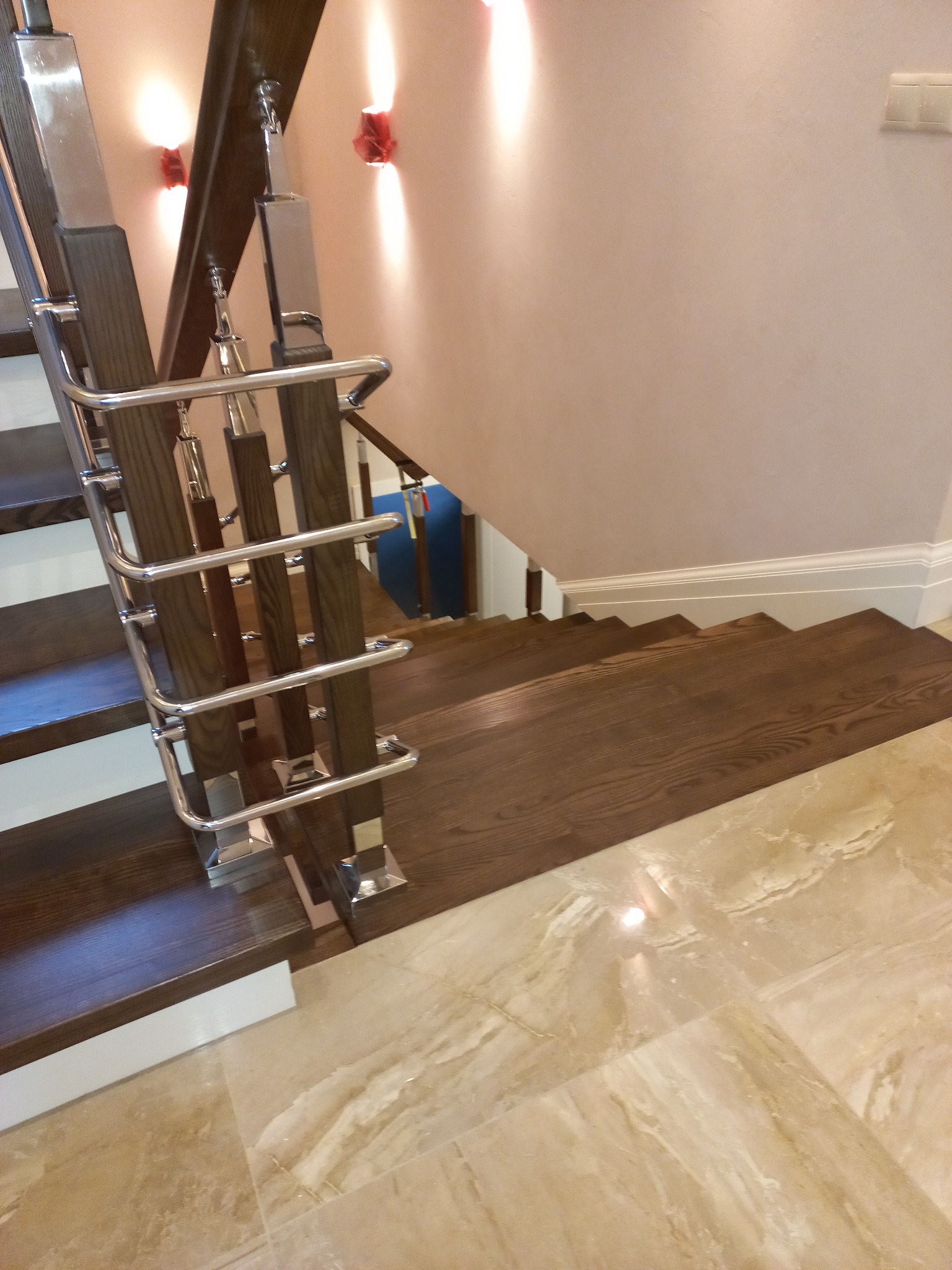 Nowoczesne schody z ciemnego drewna i metalowymi balustradami na marmurowej podłodze, oświetlone kinkietami z czerwonymi abażurami.