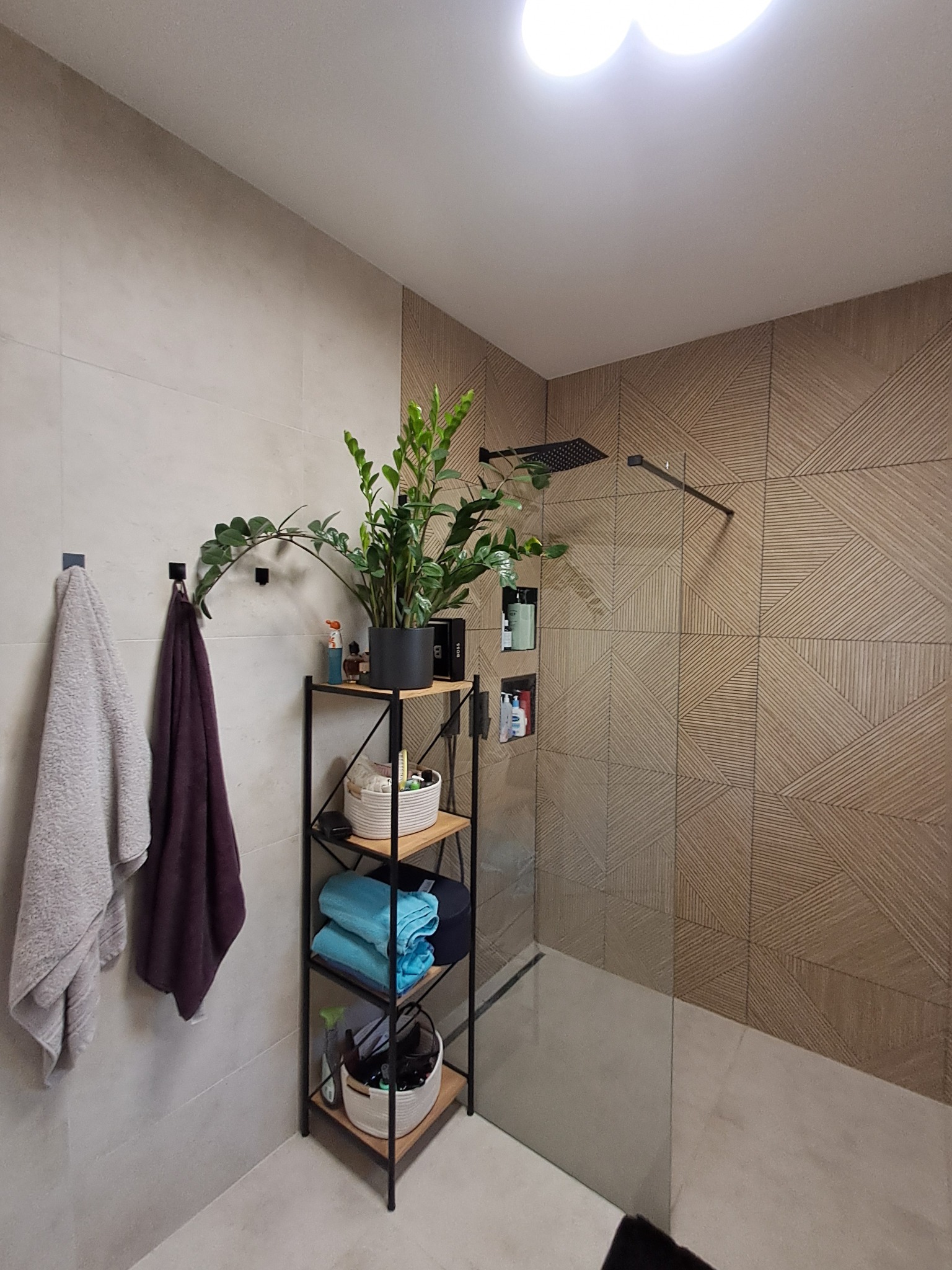 Nowoczesna łazienka z kabiną prysznicową, wykończona płytkami w geometryczny wzór, z rośliną doniczkową na metalowym regale i ręcznikami na wieszakach.