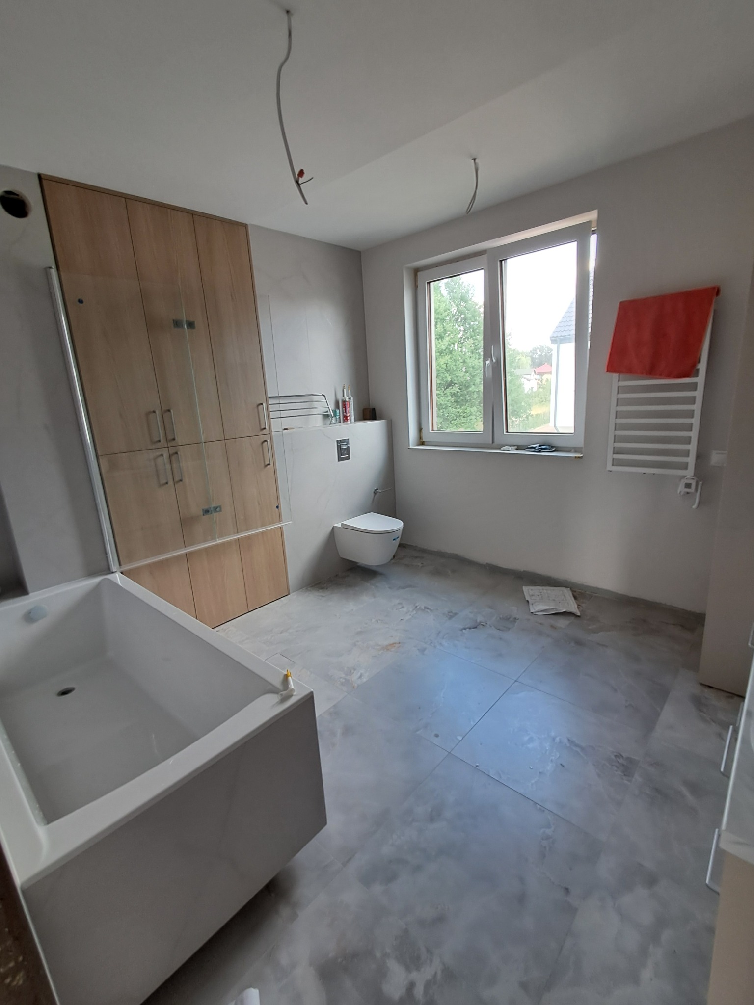 Łazienka w trakcie wykończenia: położone szare płytki imitujące marmur, biała wanna, zabudowa meblowa z jasnego drewna, wisząca toaleta i grzejnik drabinkowy z pomarańczowym ręcznikiem.