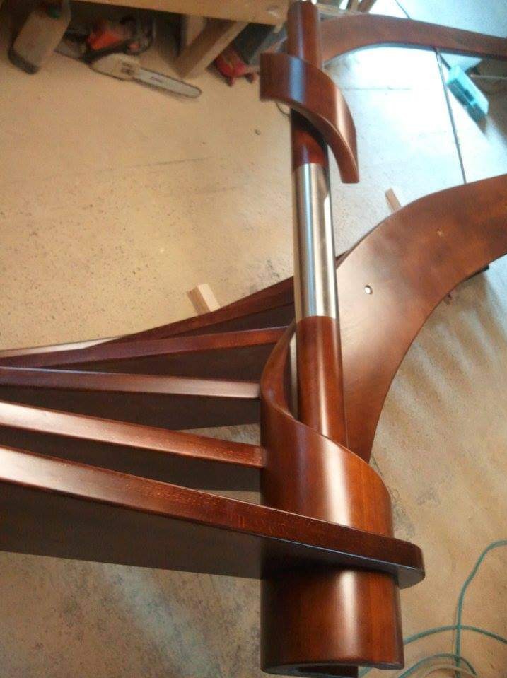 Fragment spiralnych schodów z ciemnego drewna z centralnym słupem z elementami drewnianymi i metalową rurą, widoczne narzędzia w tle.