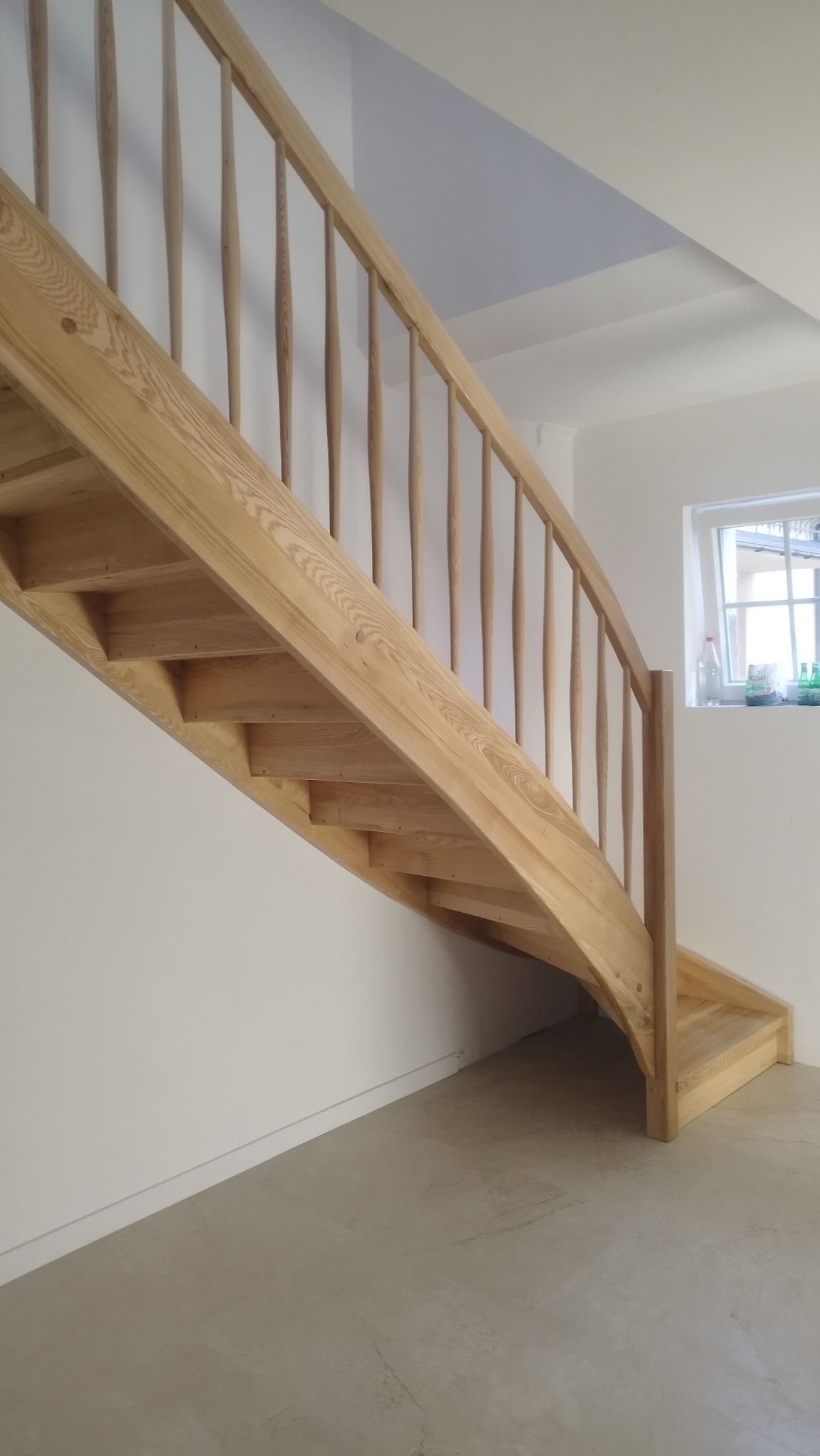 Jasne, drewniane schody z balustradą o toczonych szczeblach, widziane z dołu. Naturalne wykończenie drewna, minimalistyczne wnętrze z oknem w tle.