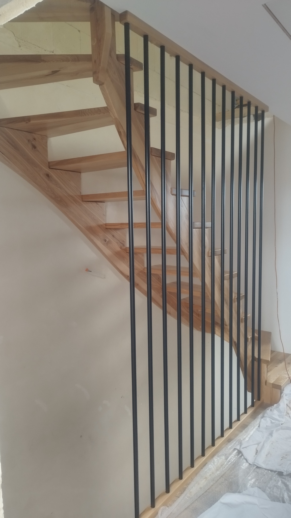 Jasne, drewniane schody z czarnymi, metalowymi prętami balustrady. Widok z dołu, ukazujący detale konstrukcji i kontrast materiałów. Minimalistyczny design.