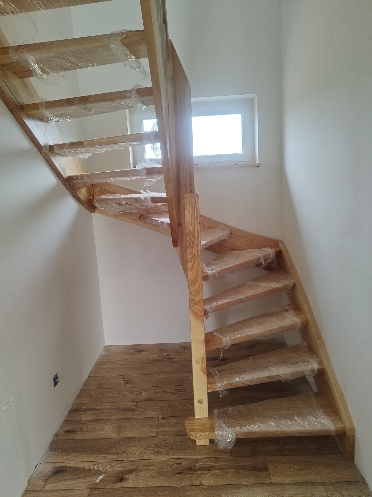 Jasne, drewniane schody w trakcie montażu, zabezpieczone folią, z widocznym fragmentem drewnianej podłogi i okna. Ujęcie z dołu, podkreślające konstrukcję.