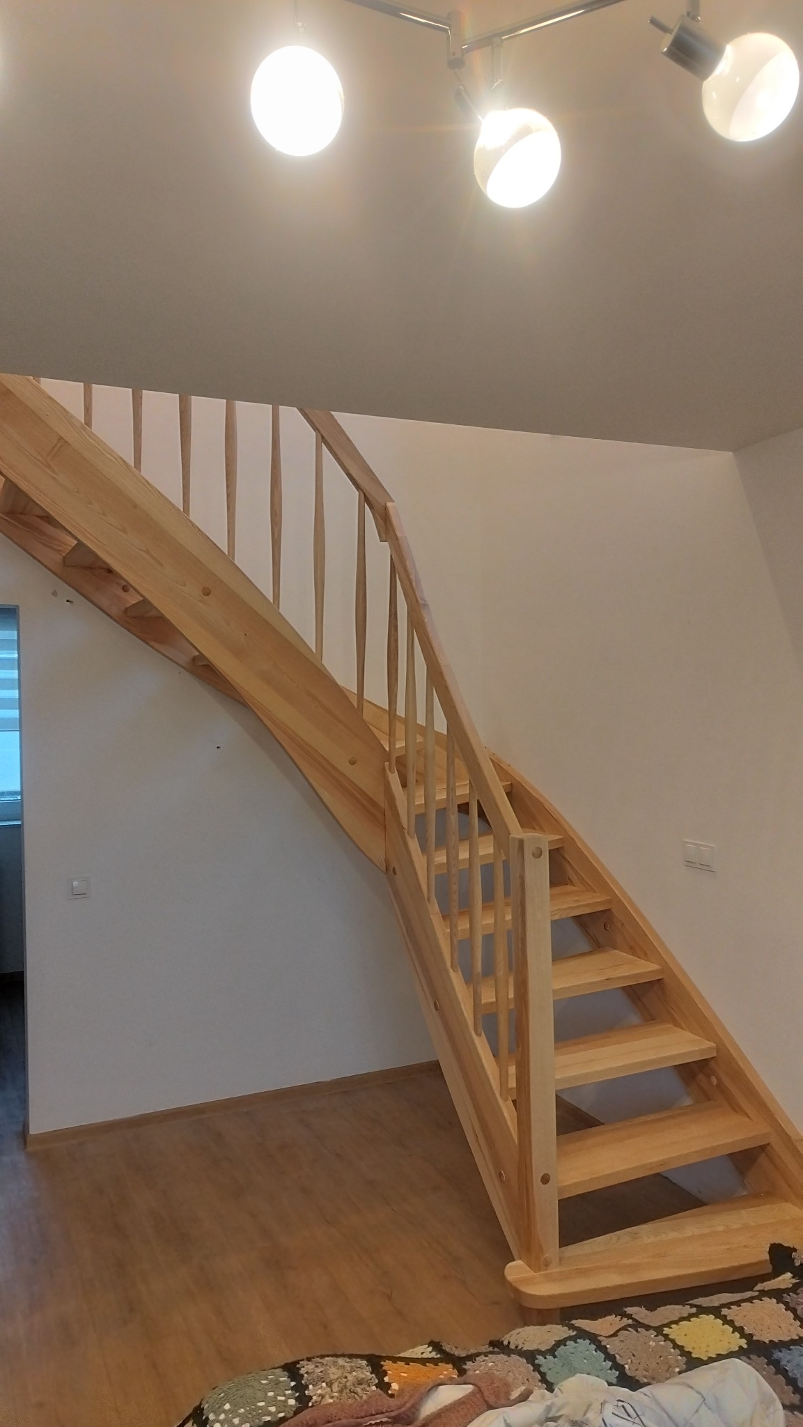 Jasne, drewniane schody z balustradą w jasnym wnętrzu. Widoczne stopnie i poręcze. U dołu schodów leży pled. Oświetlenie sufitowe z trzema punktami świetlnymi.