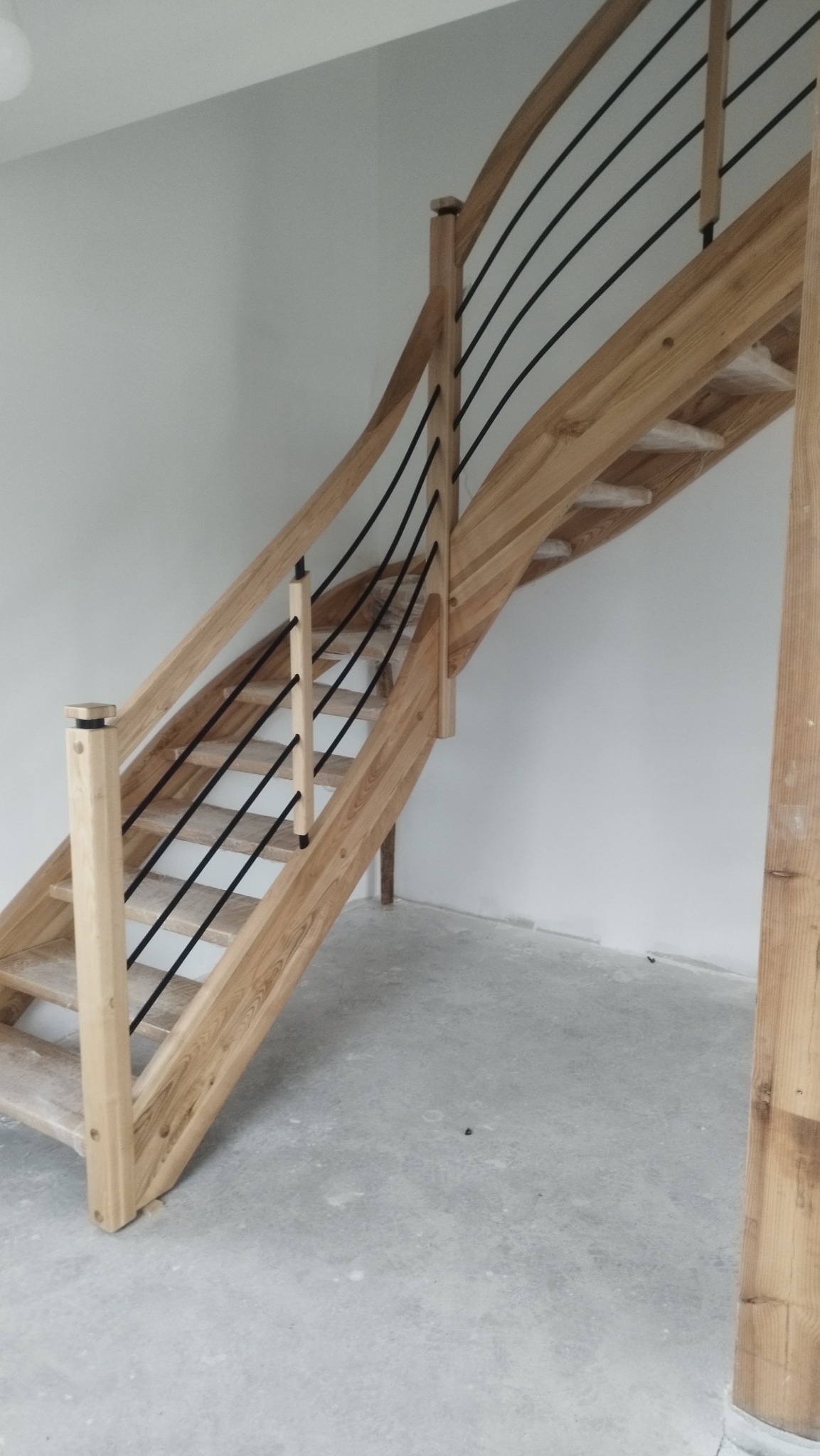 Jasne, drewniane schody z czarnymi, metalowymi prętami balustrady, widziane z dołu, w surowym wnętrzu. Elementy schodów zabezpieczone folią.