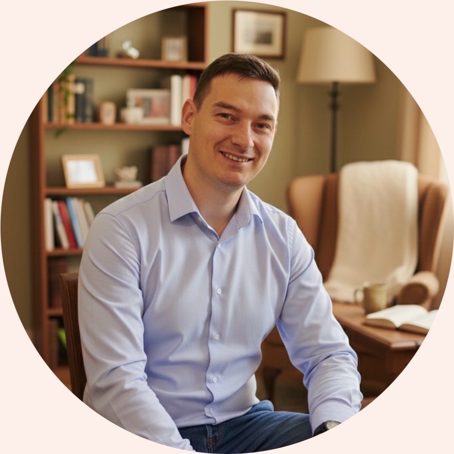 Uśmiechnięty mężczyzna w błękitnej koszuli siedzi na krześle, w tle regał z książkami, lampa i fotel. Portret w ciepłych barwach, w okrągłej ramce.