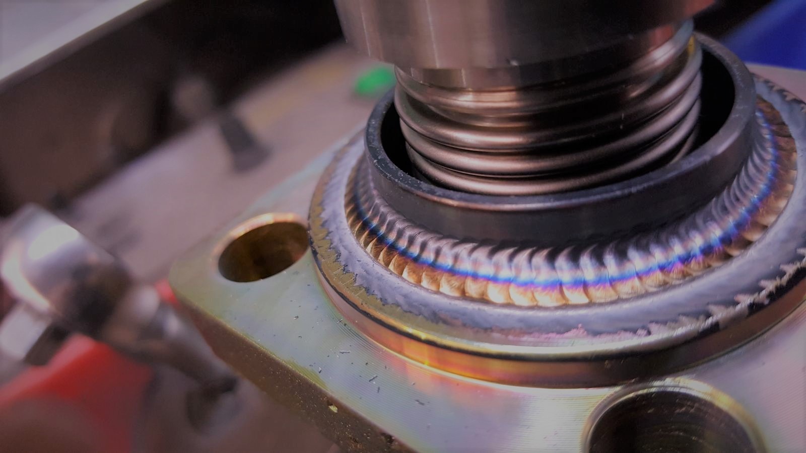 Zbliżenie na precyzyjny spaw na metalowej konstrukcji, z widocznymi tęczowymi przebarwieniami termicznymi i spiralnym elementem wewnątrz, uchwycone pod kątem.