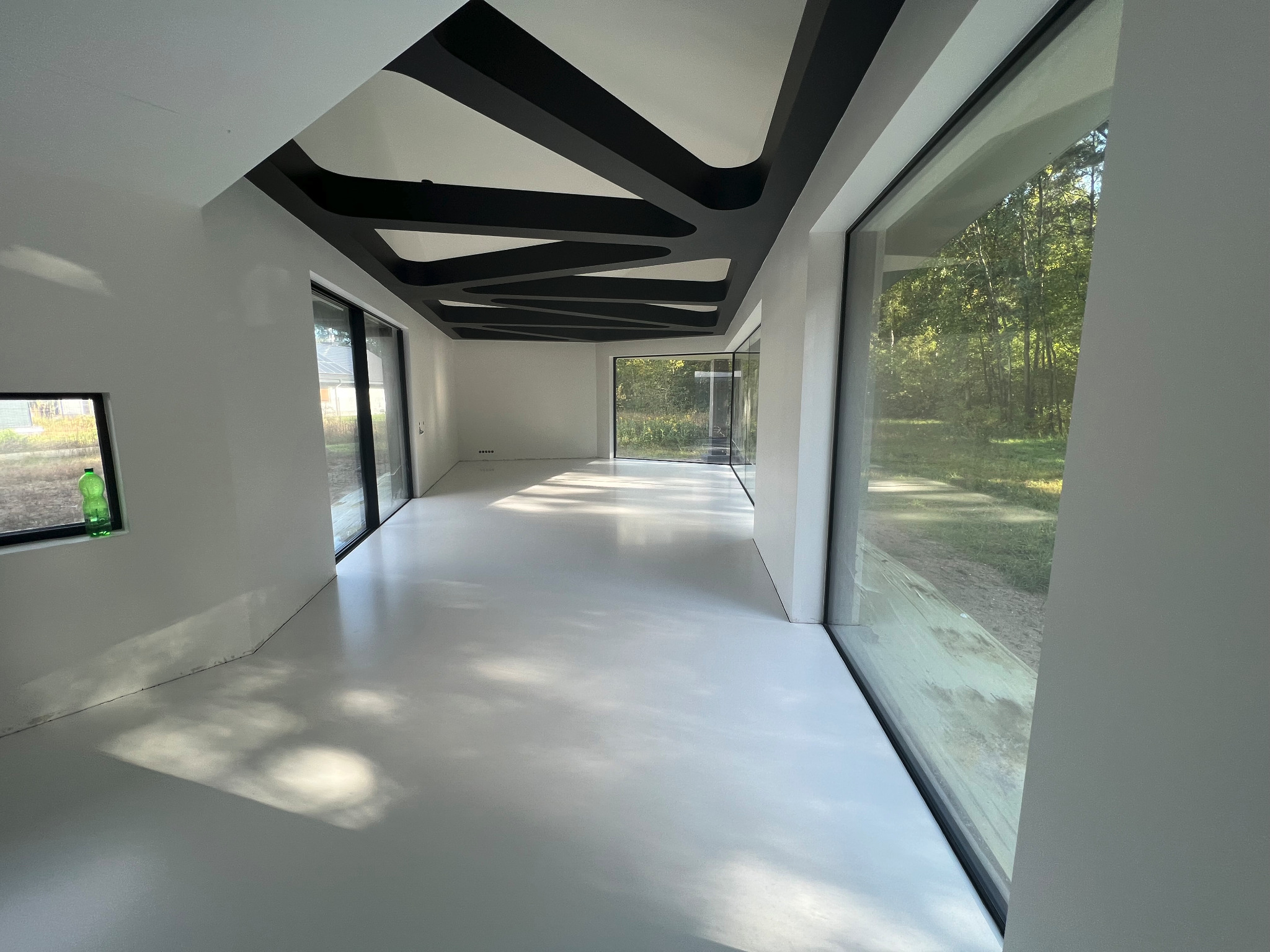 Wnętrze nowoczesnego domu z białą, błyszczącą podłogą, czarnymi, geometrycznymi elementami dekoracyjnymi na suficie i dużymi oknami z widokiem na zieleń.