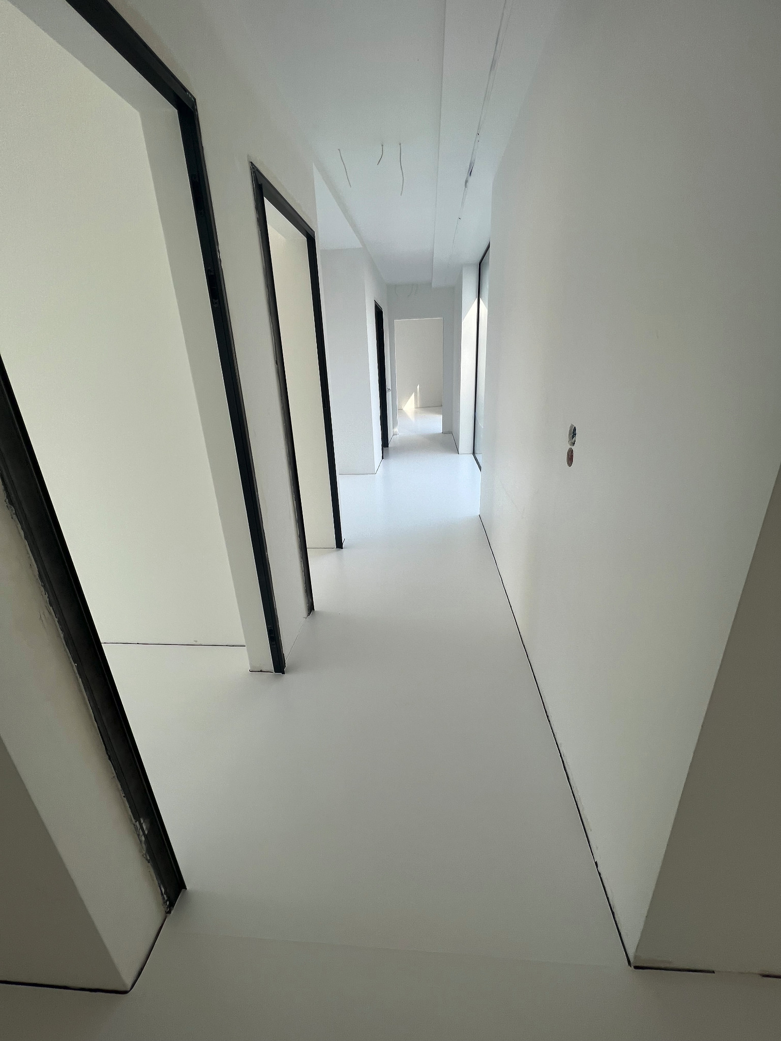 Biały korytarz z czarnymi futrynami drzwiowymi i jednolitą, jasną posadzką.