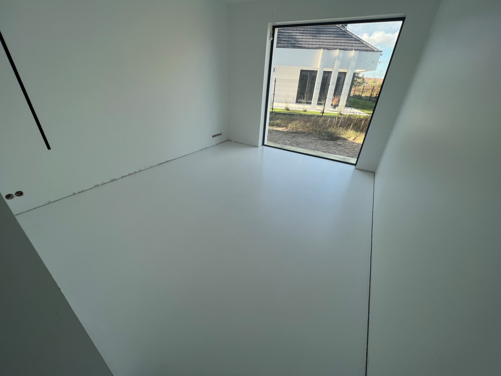 Jasne, jednolite wykończenie podłogi w pustym pomieszczeniu z dużym oknem na dom z szarą dachówką.