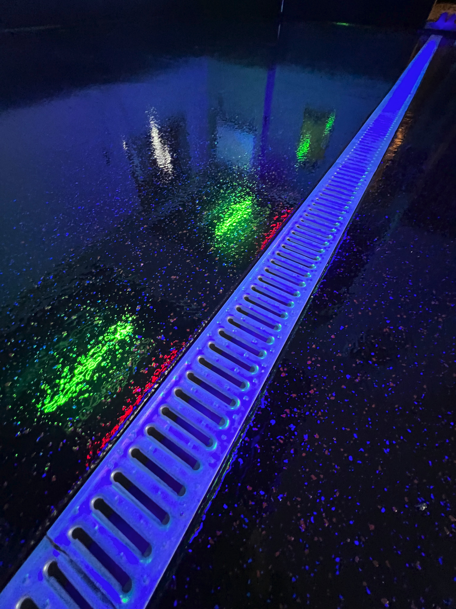 Błyszcząca, czarna posadzka z brokatem podświetlona niebieskim światłem UV, z liniowym odpływem podłogowym.