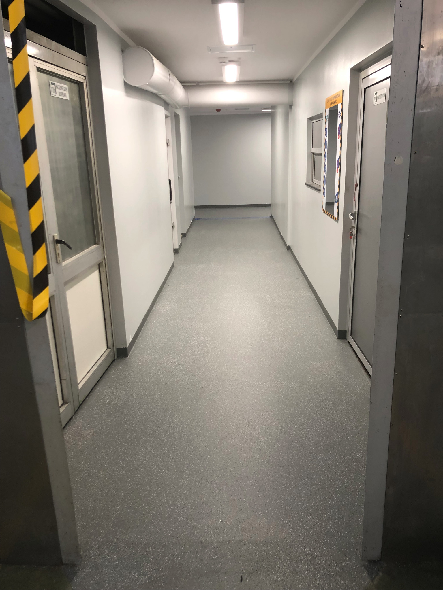 Szary korytarz z posadzką żywiczną w budynku użyteczności publicznej, widoczne drzwi i żółto-czarne oznaczenia ostrzegawcze.