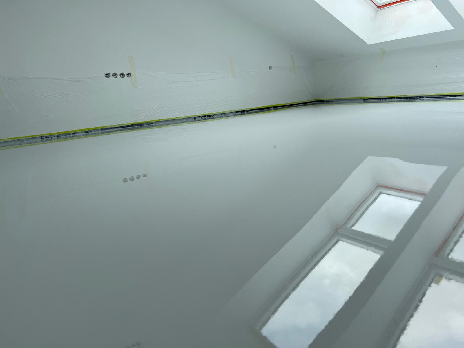 Świeżo wylana, błyszcząca, szara posadzka żywiczna w pomieszczeniu z oknami, zabezpieczone ściany folią malarską i taśmą. Widoczne puszki elektryczne w ścianie.
