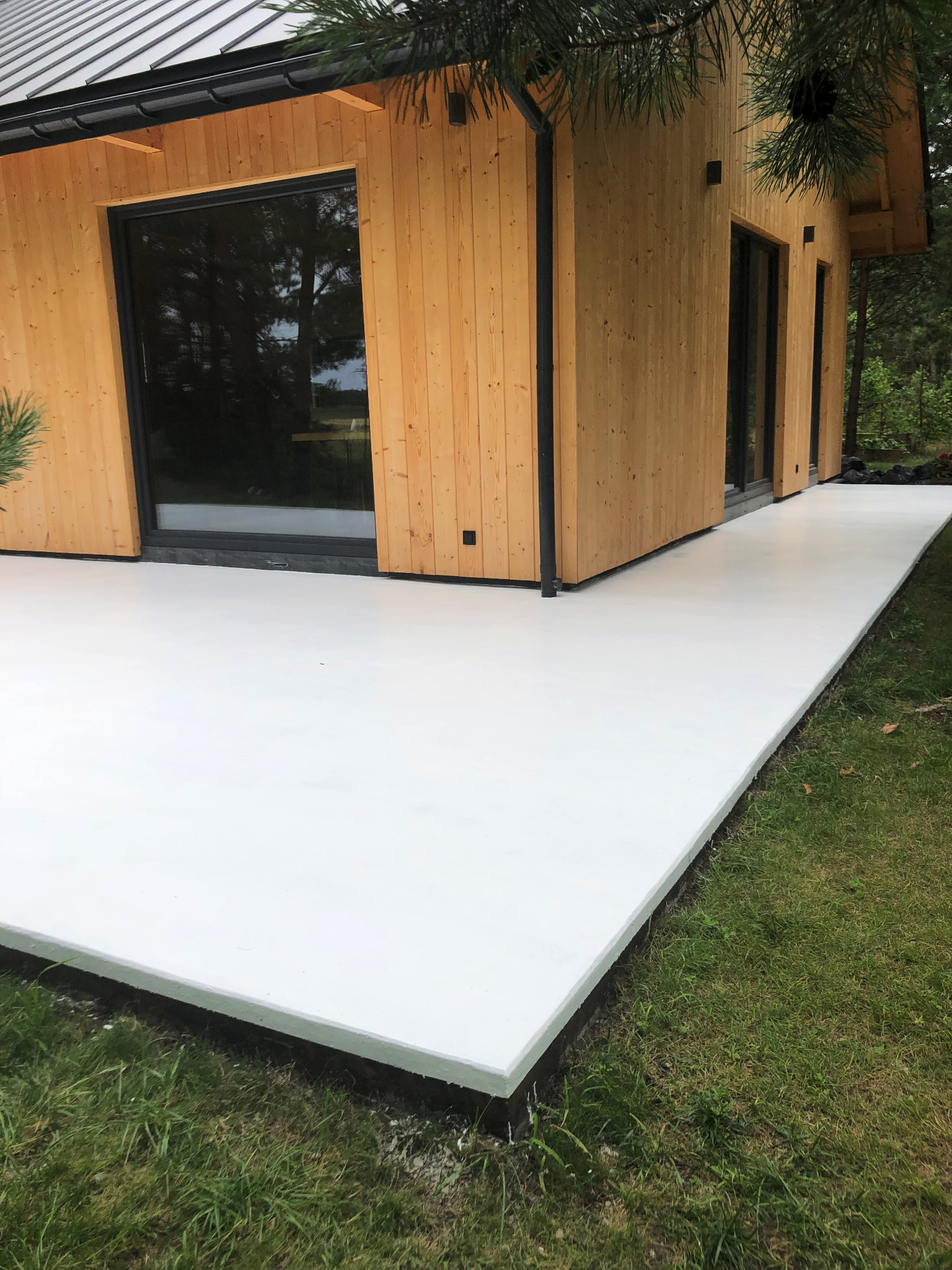 Biała, gładka posadzka na zewnątrz drewnianego budynku z dużymi oknami, otoczona zielonym trawnikiem i drzewami.