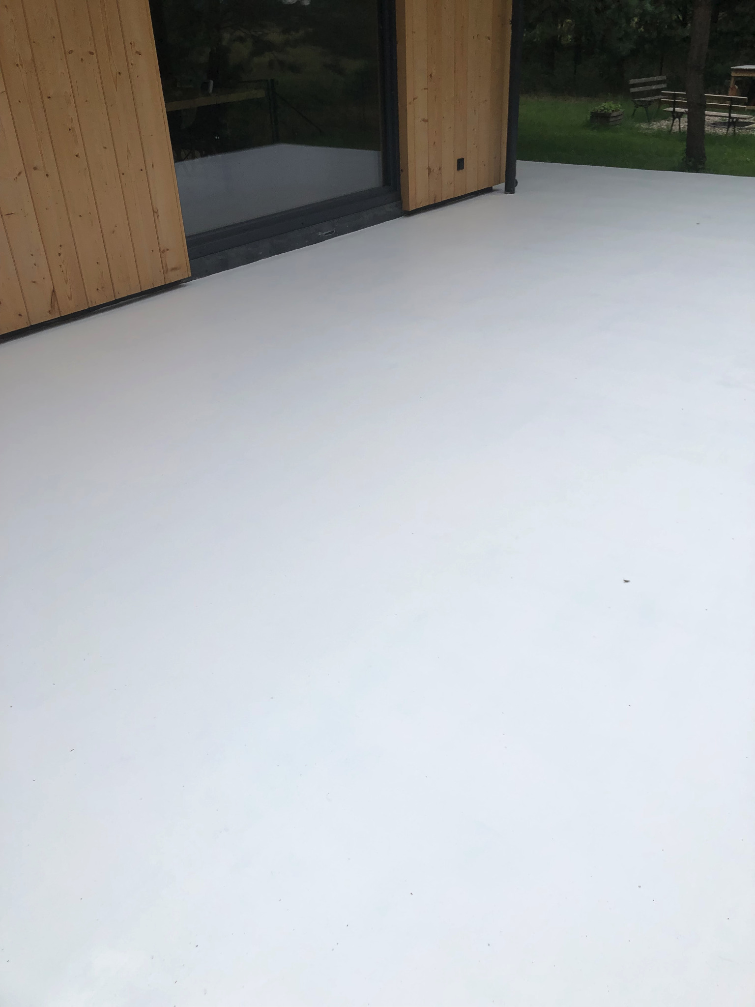 Jasnoszara, gładka powierzchnia tarasu z widocznymi drewnianymi drzwiami przesuwnymi i ogrodem w tle.