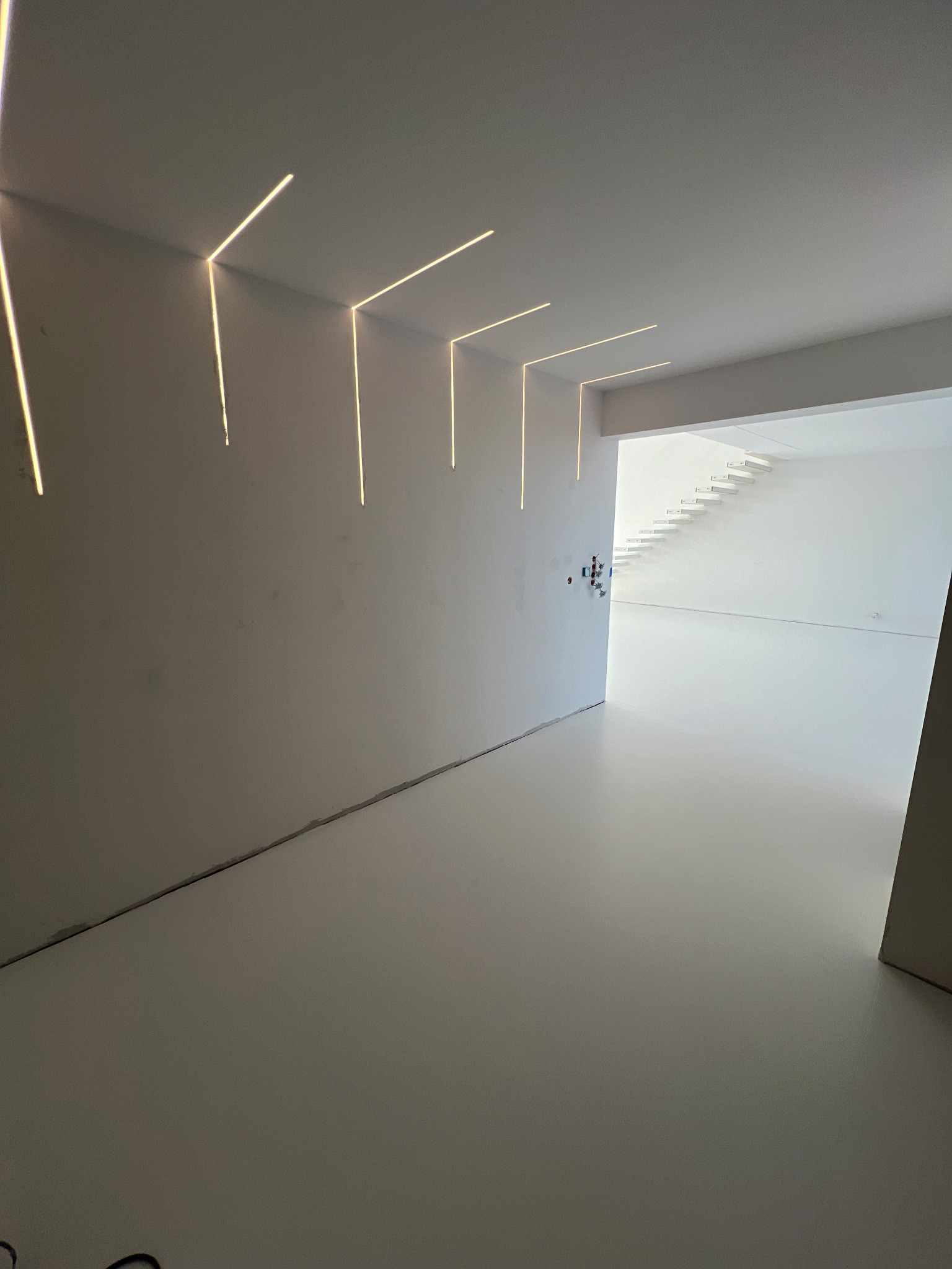 Minimalistyczne wnętrze z jasną, jednolitą posadzką i liniowym oświetleniem LED w ścianie, prowadzącym wzrok w stronę białych schodów w głębi.
