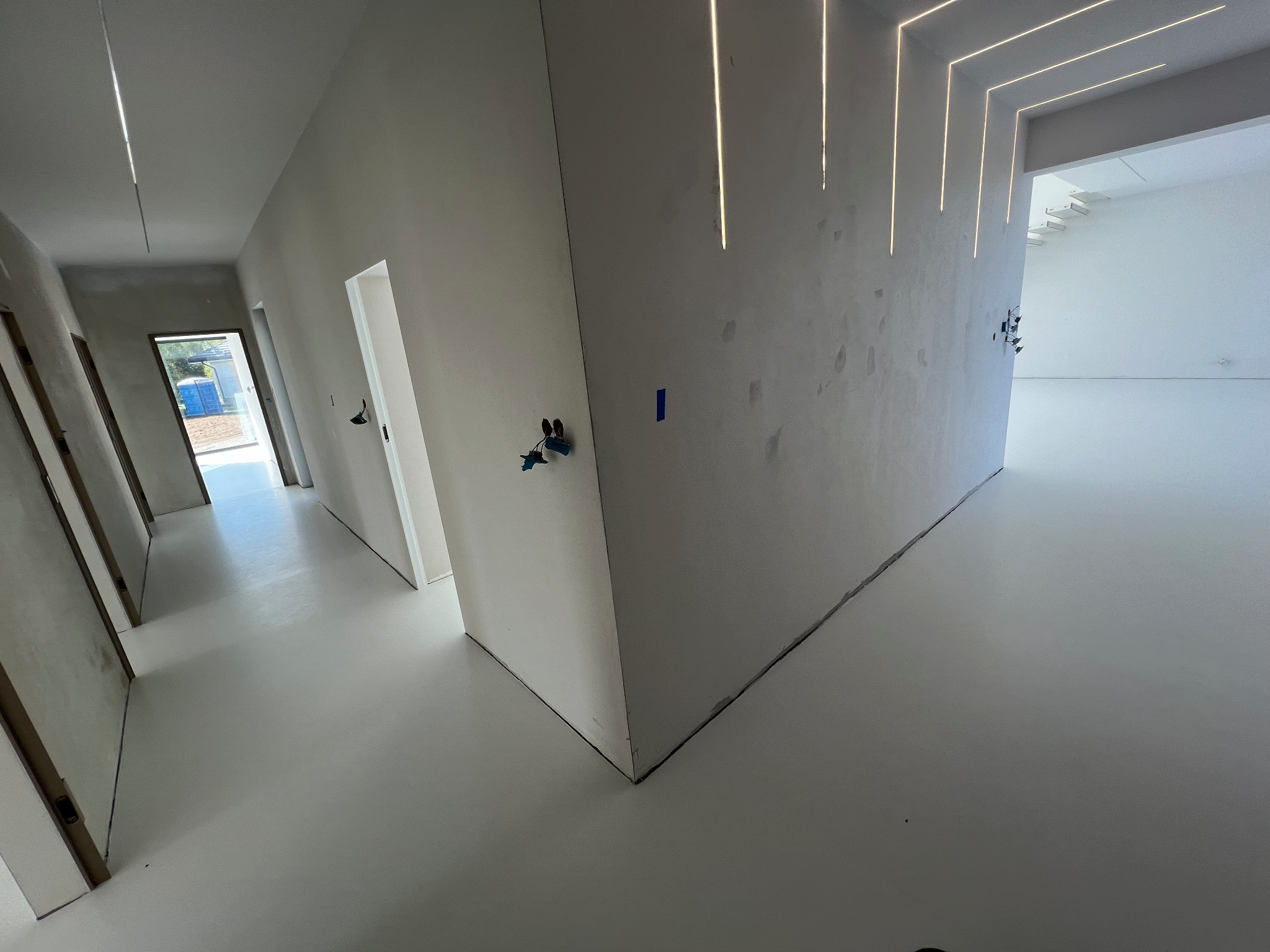 Jasnoszara posadzka żywiczna w korytarzu z oświetleniem LED w suficie i ścianach, widoczne niedokończone prace wykończeniowe.