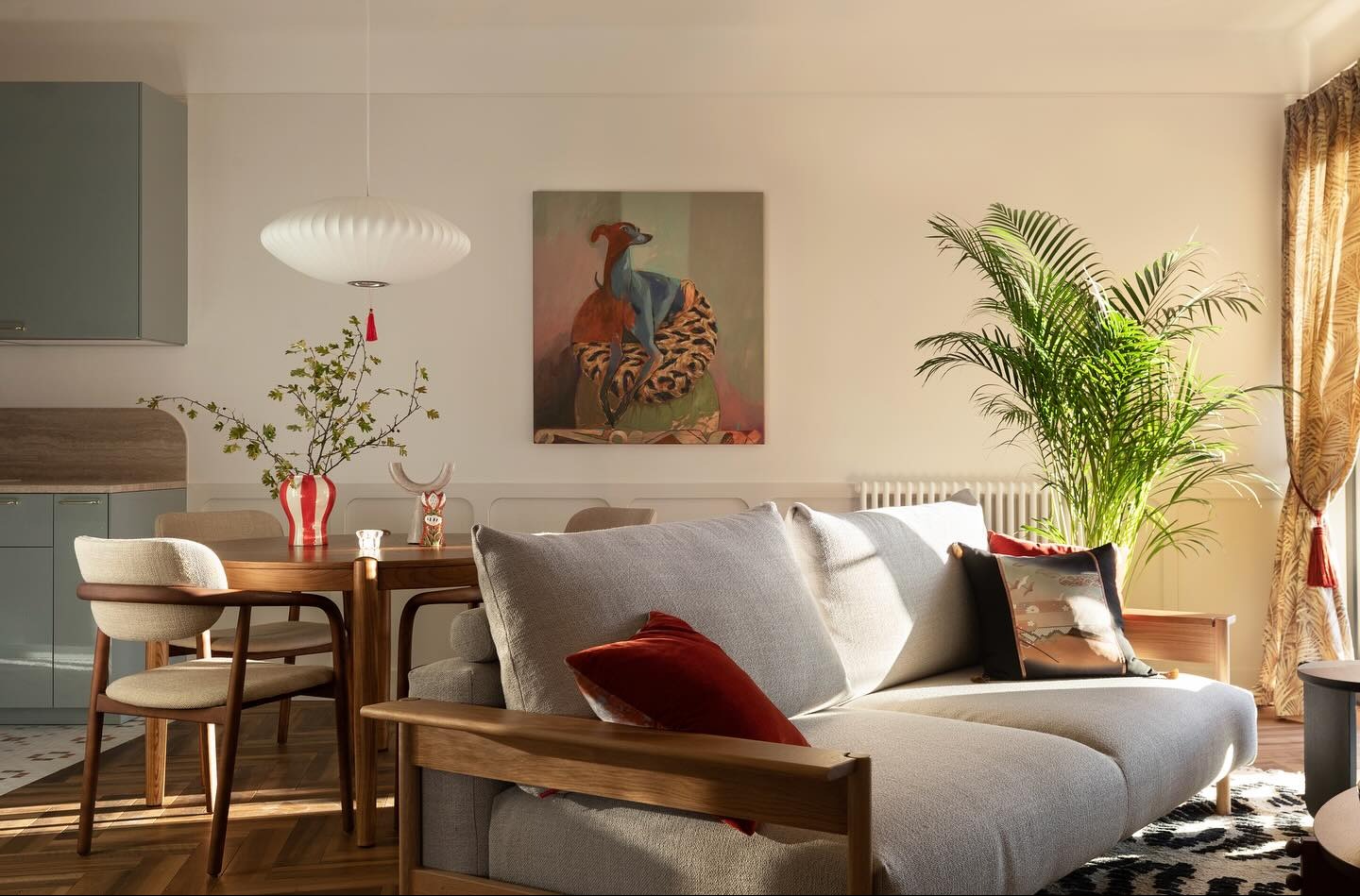 Jasne wnętrze mieszkania z drewnianym stołem, szarą sofą z poduszkami, obrazem psa i palmą, oświetlone wiszącą lampą z frędzlami.