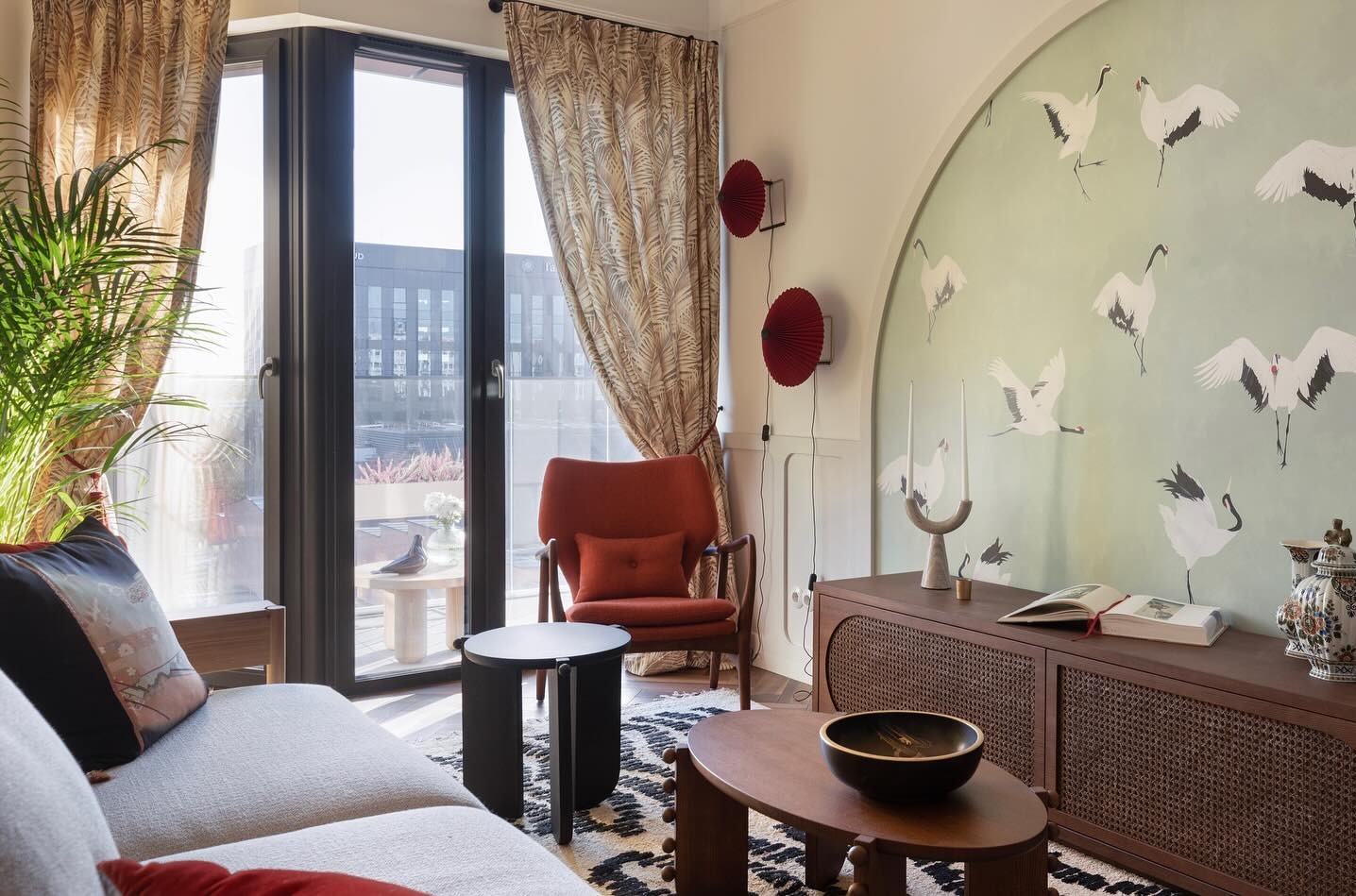 Jasne wnętrze pokoju dziennego z akcentami w stylu japońskim: tapeta z żurawiami, czerwone wachlarze na ścianie, brązowe meble z rattanowymi frontami, okno balkonowe z widokiem na miasto.