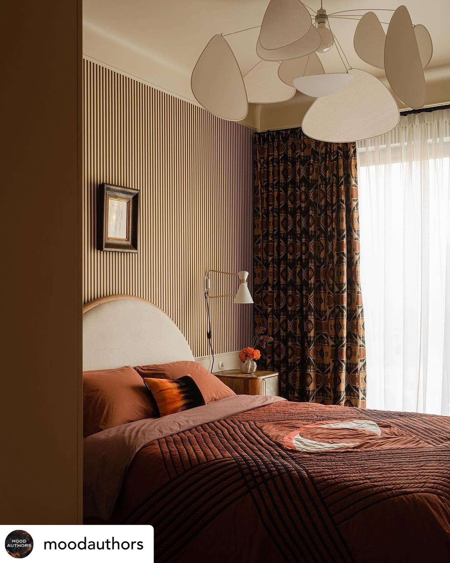 Sypialnia w odcieniach brązu i beżu z łóżkiem z pikowaną narzutą w geometryczne wzory, drewnianą ścianą w pionowe pasy, oryginalną lampą sufitową i wzorzystymi zasłonami.