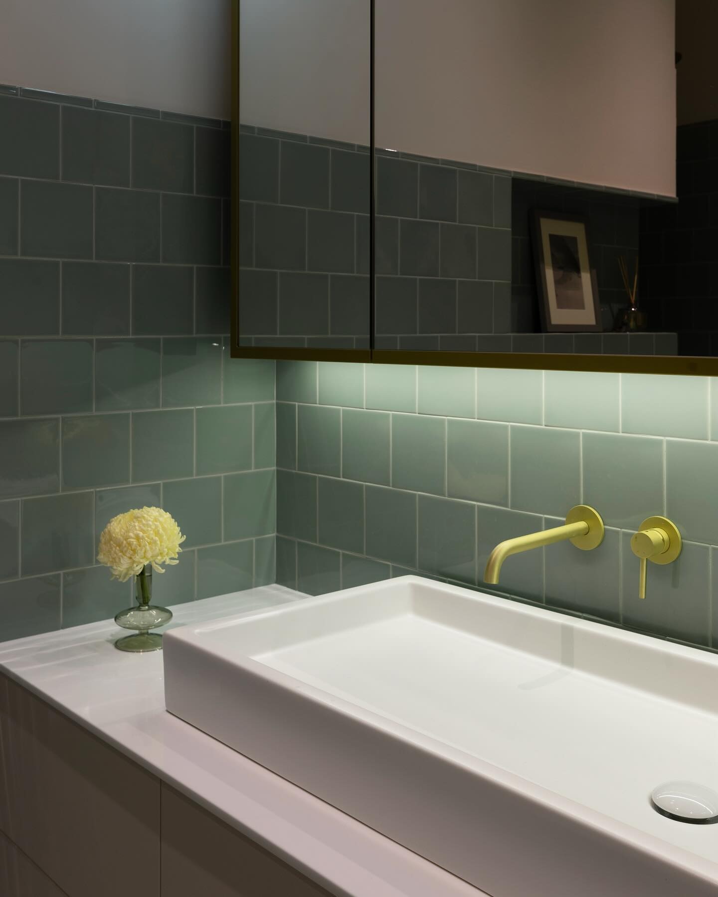 Nowoczesna łazienka z kwadratową umywalką nablatową, baterią w kolorze limonki na tle zielonych, kwadratowych płytek i podświetlanego lustra z szafkami.