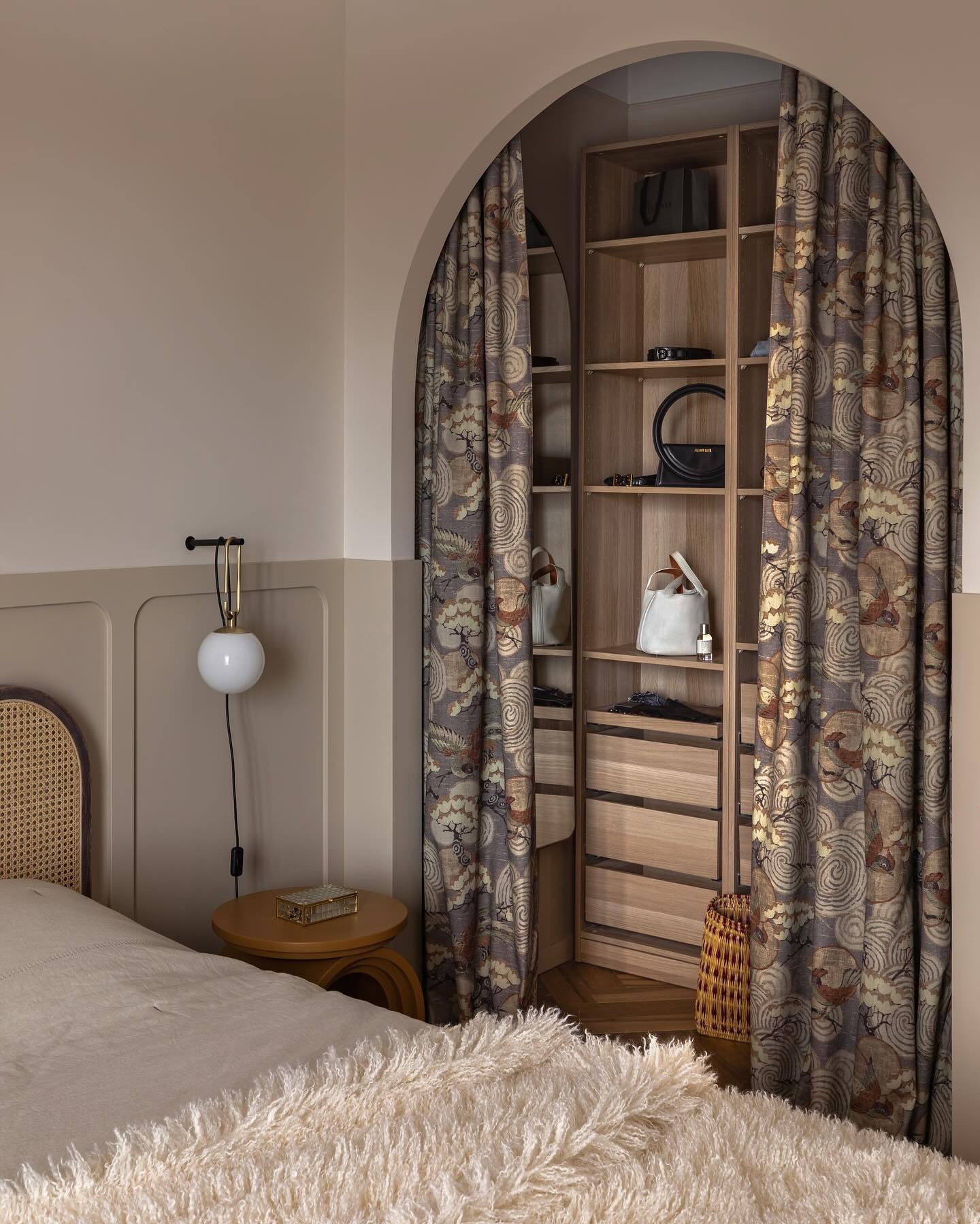 Aranżacja sypialni z otwartą garderobą oddzieloną łukiem z zasłonami w orientalny wzór, widoczny fragment łóżka z futrzaną narzutą, na stoliku nocnym szklane pudełko, na ścianie lampa kula.